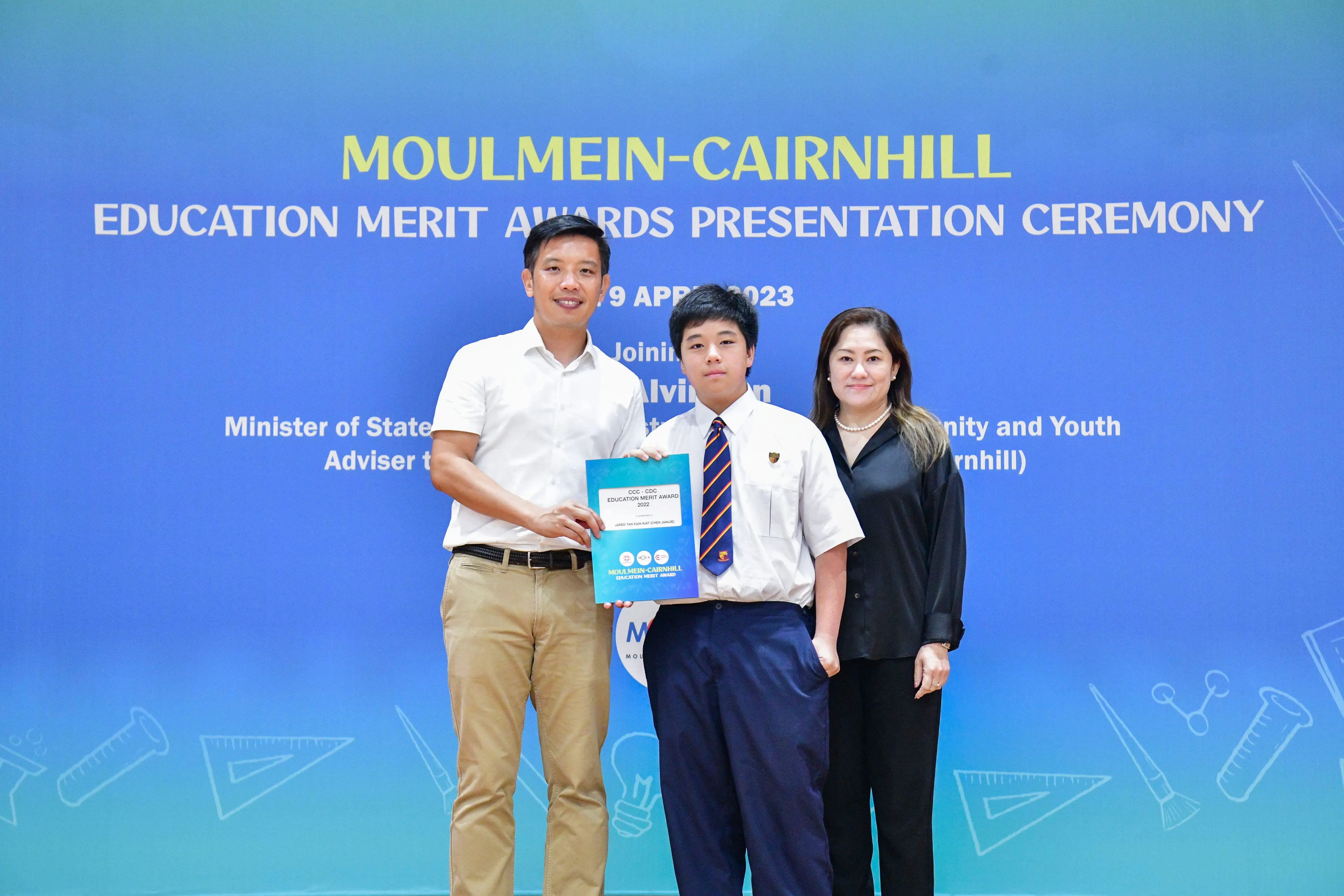 Three people pose at the Moulmein-Cairnhill Education Merit Awards Ceremony, with a student holding a blue award folder.