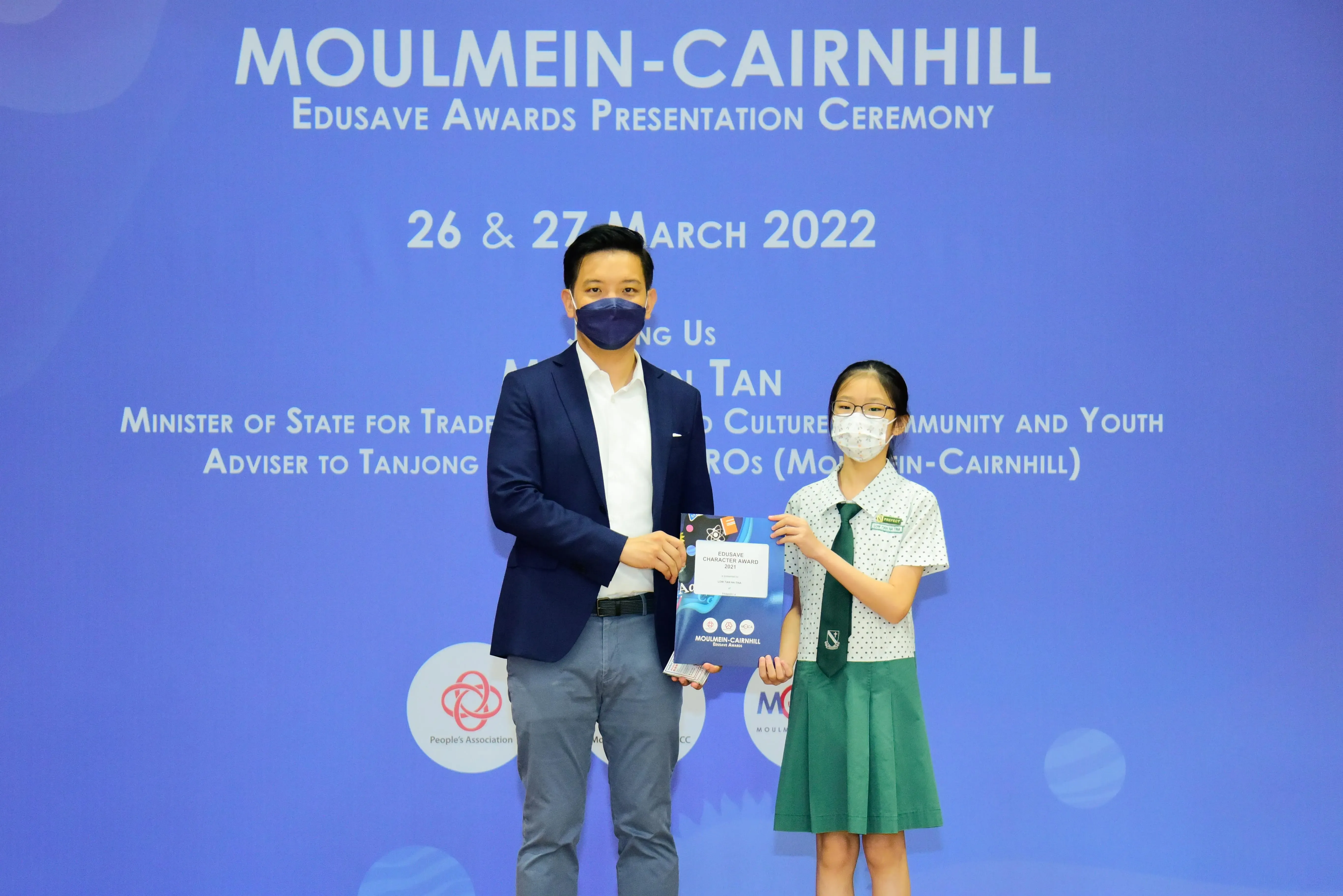 An adult and a young student in school uniform hold an Edusave award at the Moulmein-Cairnhill ceremony, March 2022.