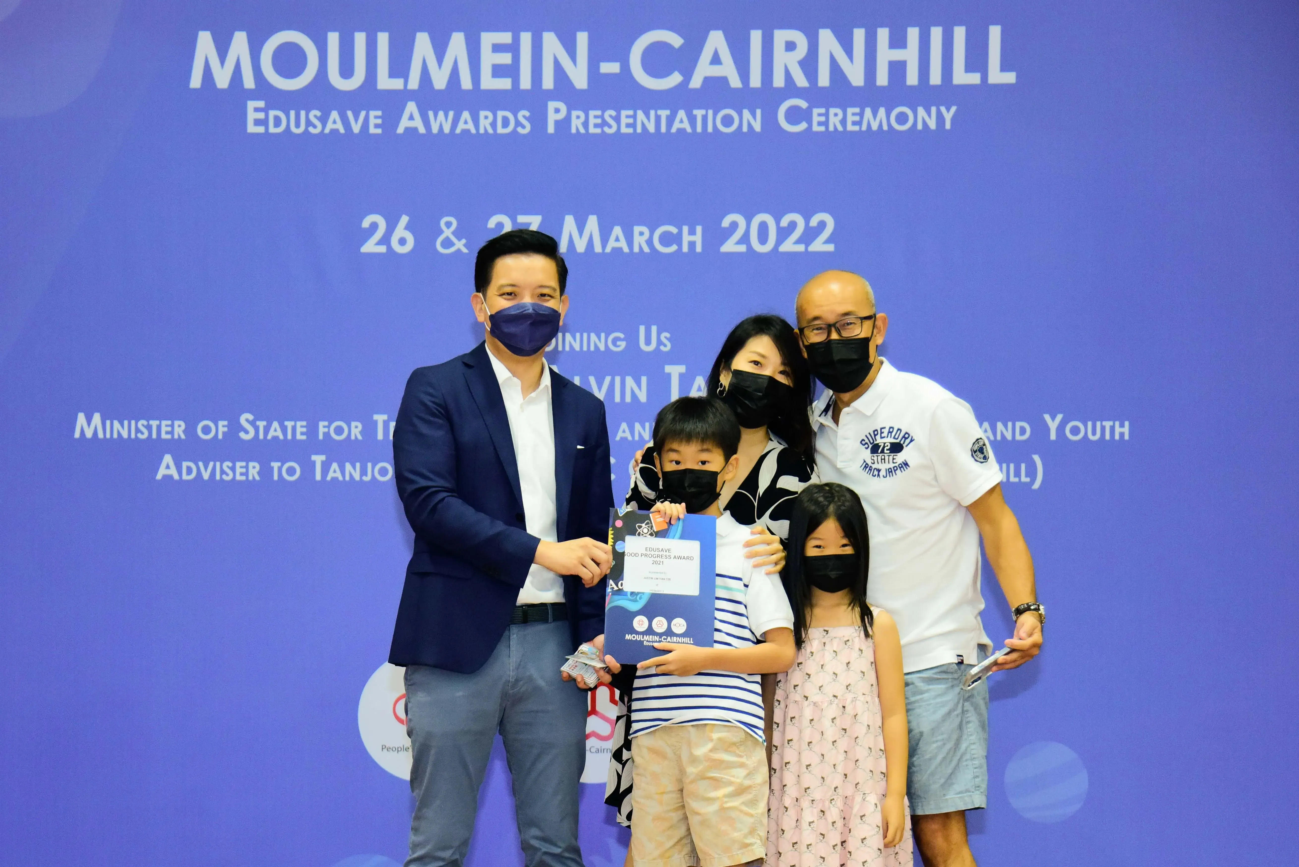 Five masked people pose at the Moulmein-Cairnhill Edusave Awards Ceremony on 26-27 March 2022, child holding award.