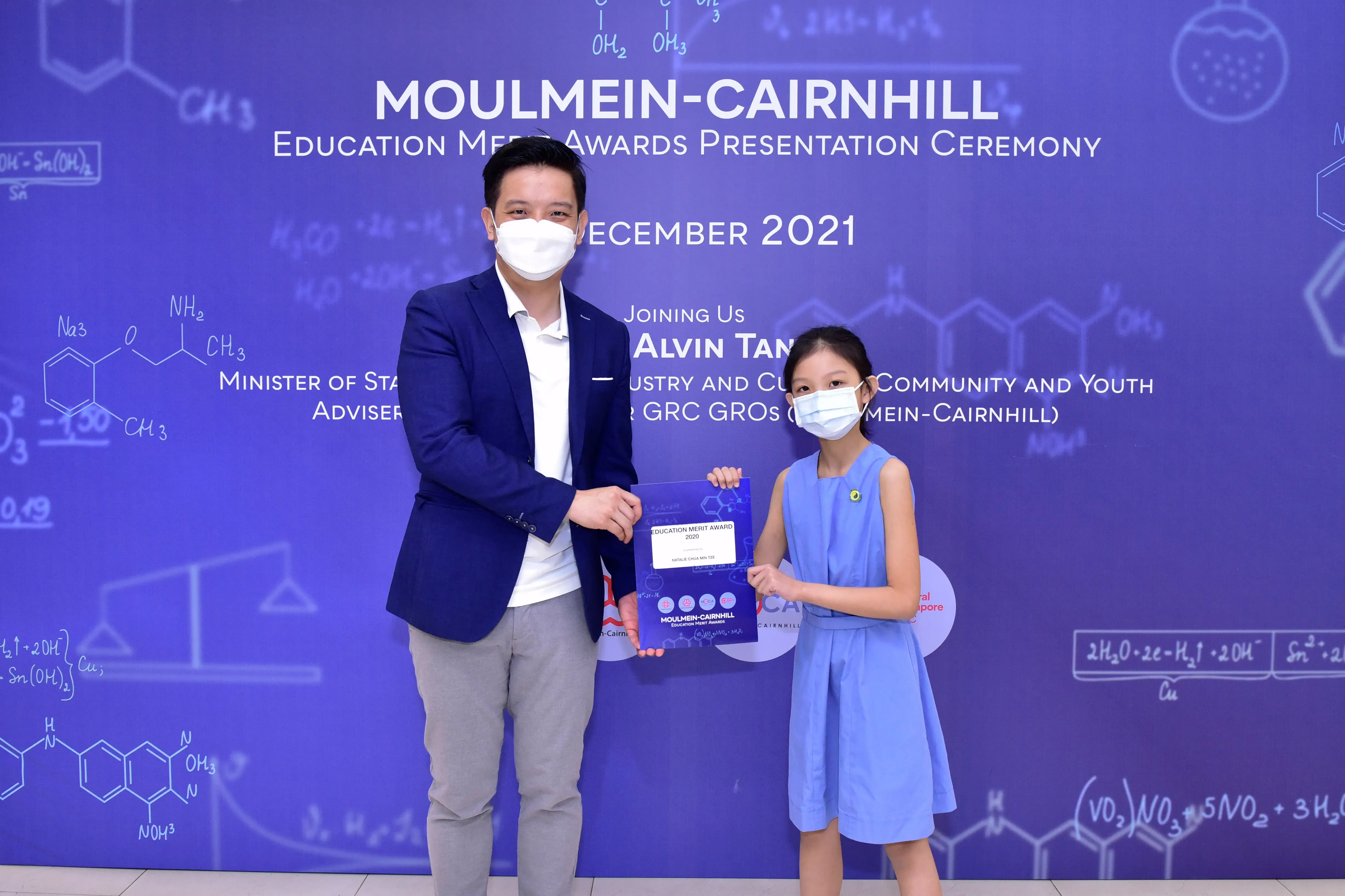 A masked man in a blazer presents an award certificate to a young girl in a blue dress at a 2021 ceremony.