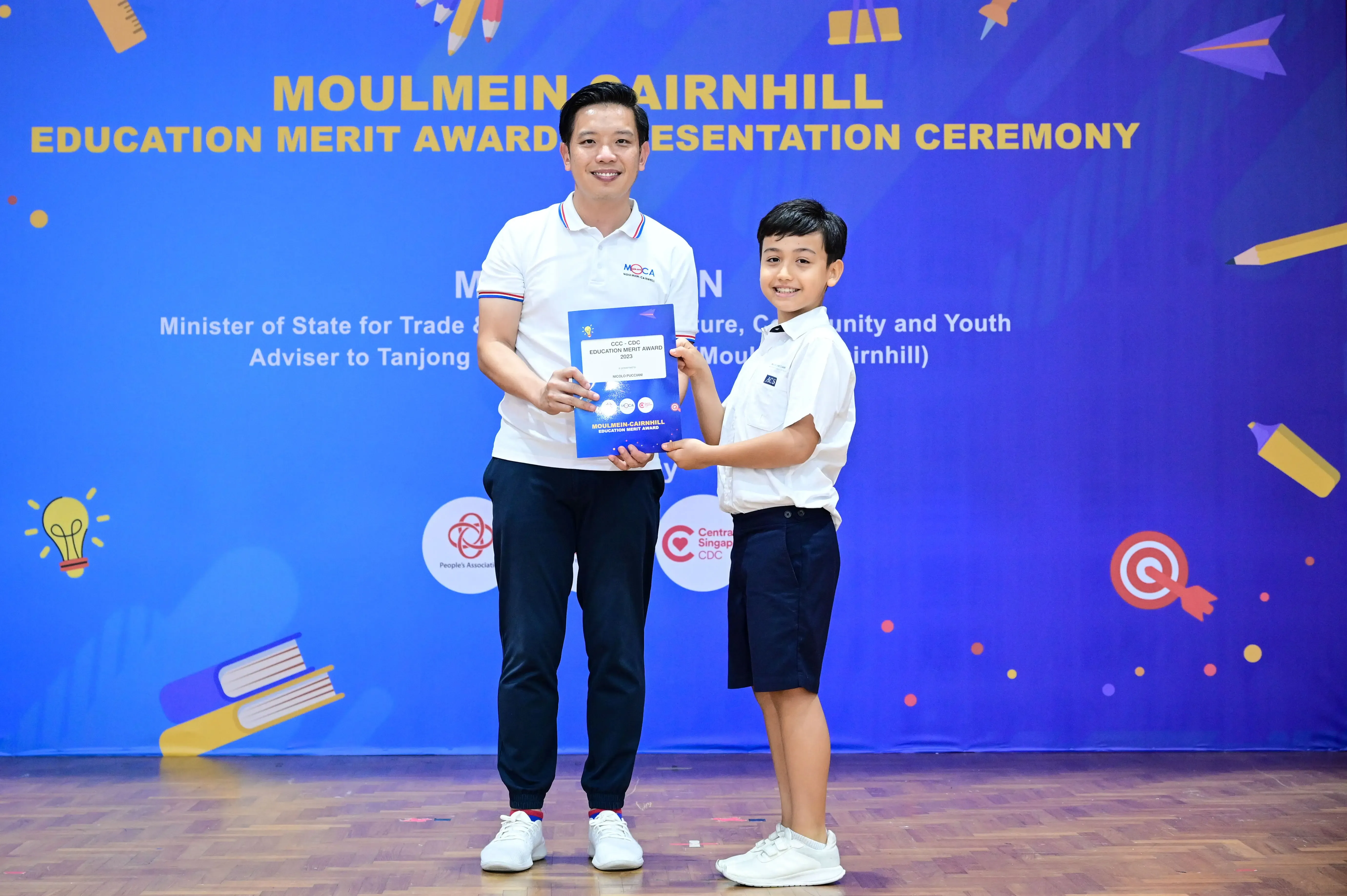 Adult and young boy holding a certificate together at the Moulmein-Cairnhill Education Merit Award Presentation Ceremony.
