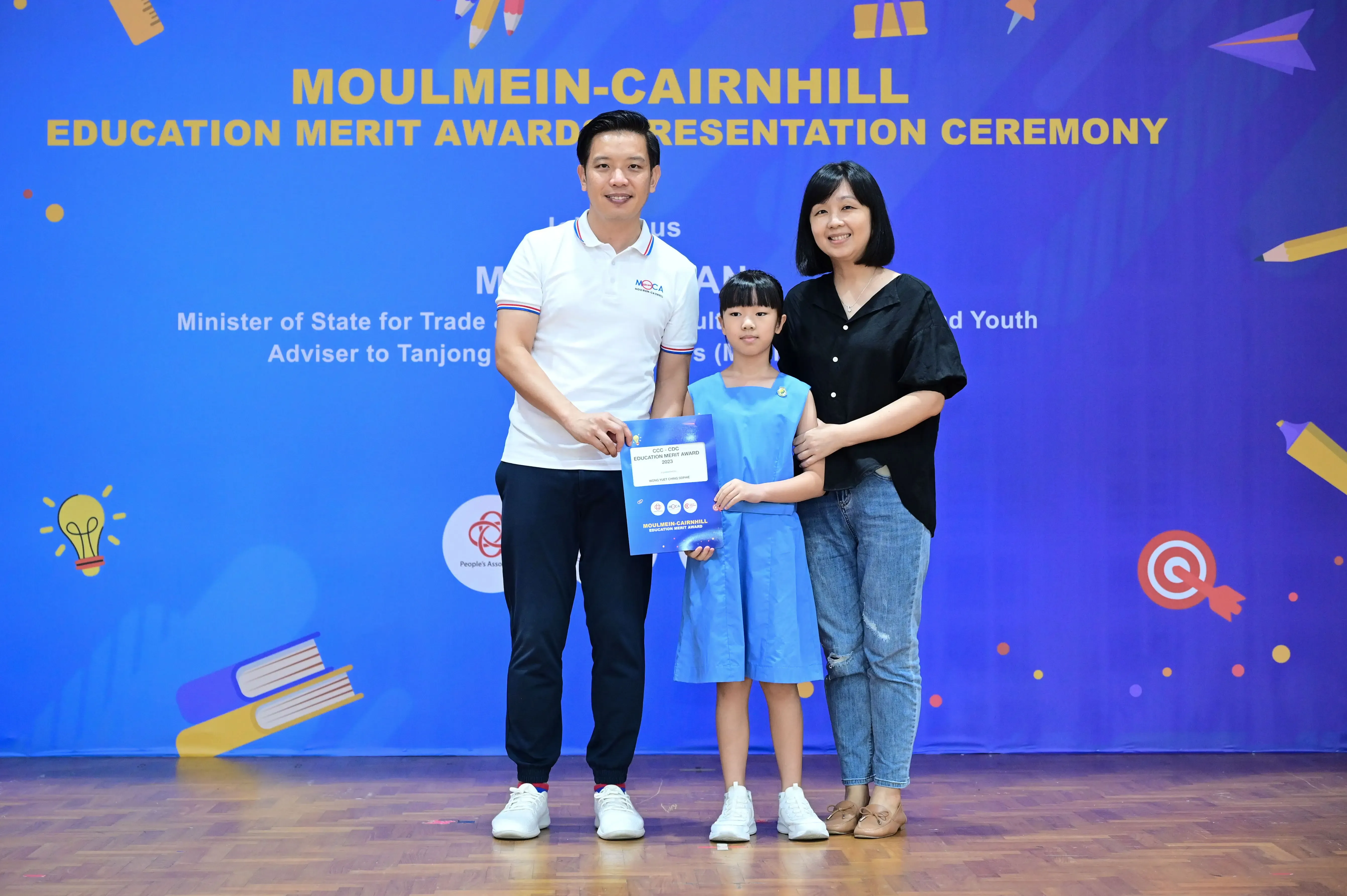 A man, young girl holding a certificate, and a woman pose at the Moulmein-Cairnhill Education Merit Award Ceremony.