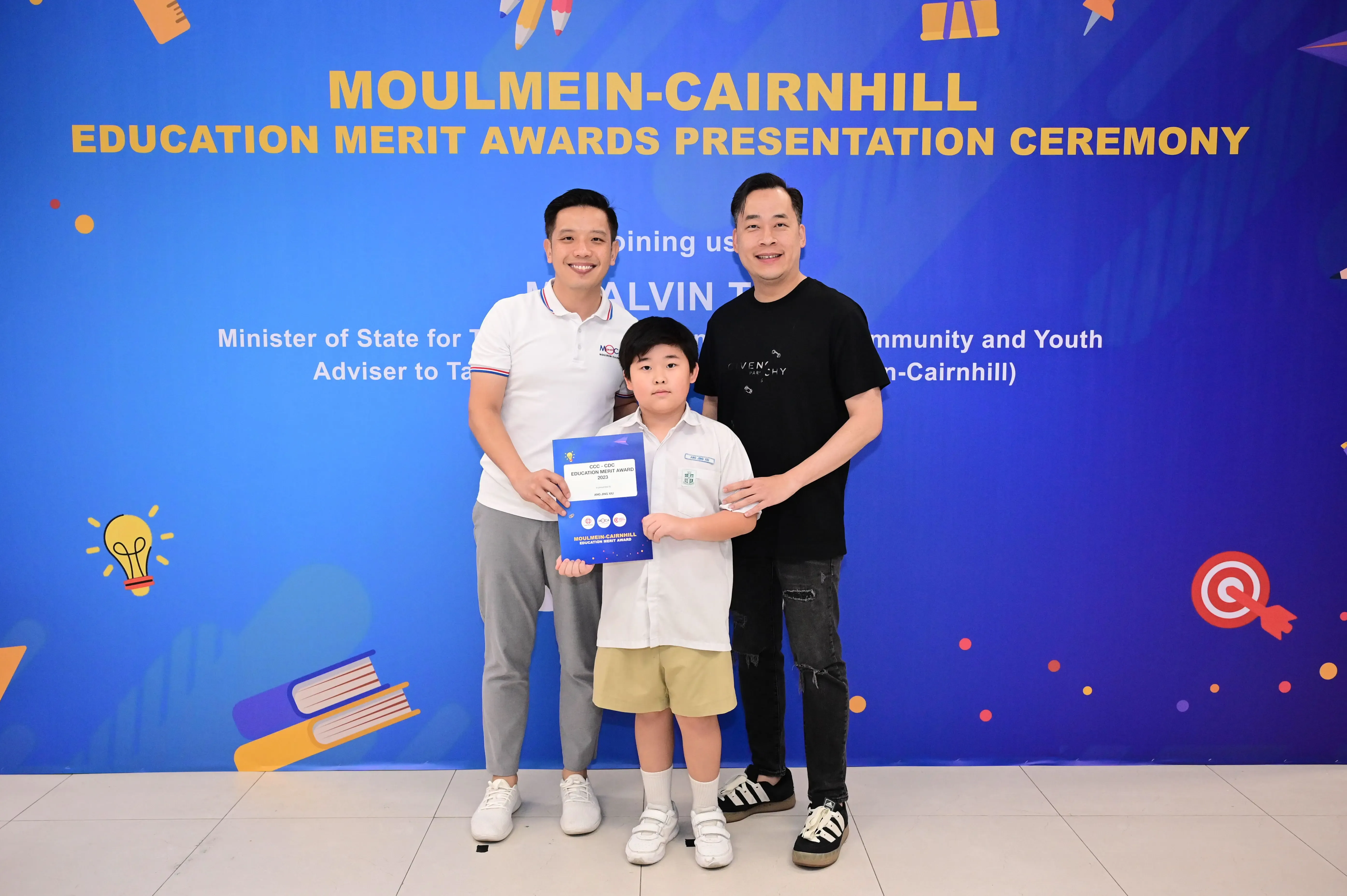 Two men and a boy holding an award certificate pose before a Moulmein-Cairnhill Education Merit Awards banner.