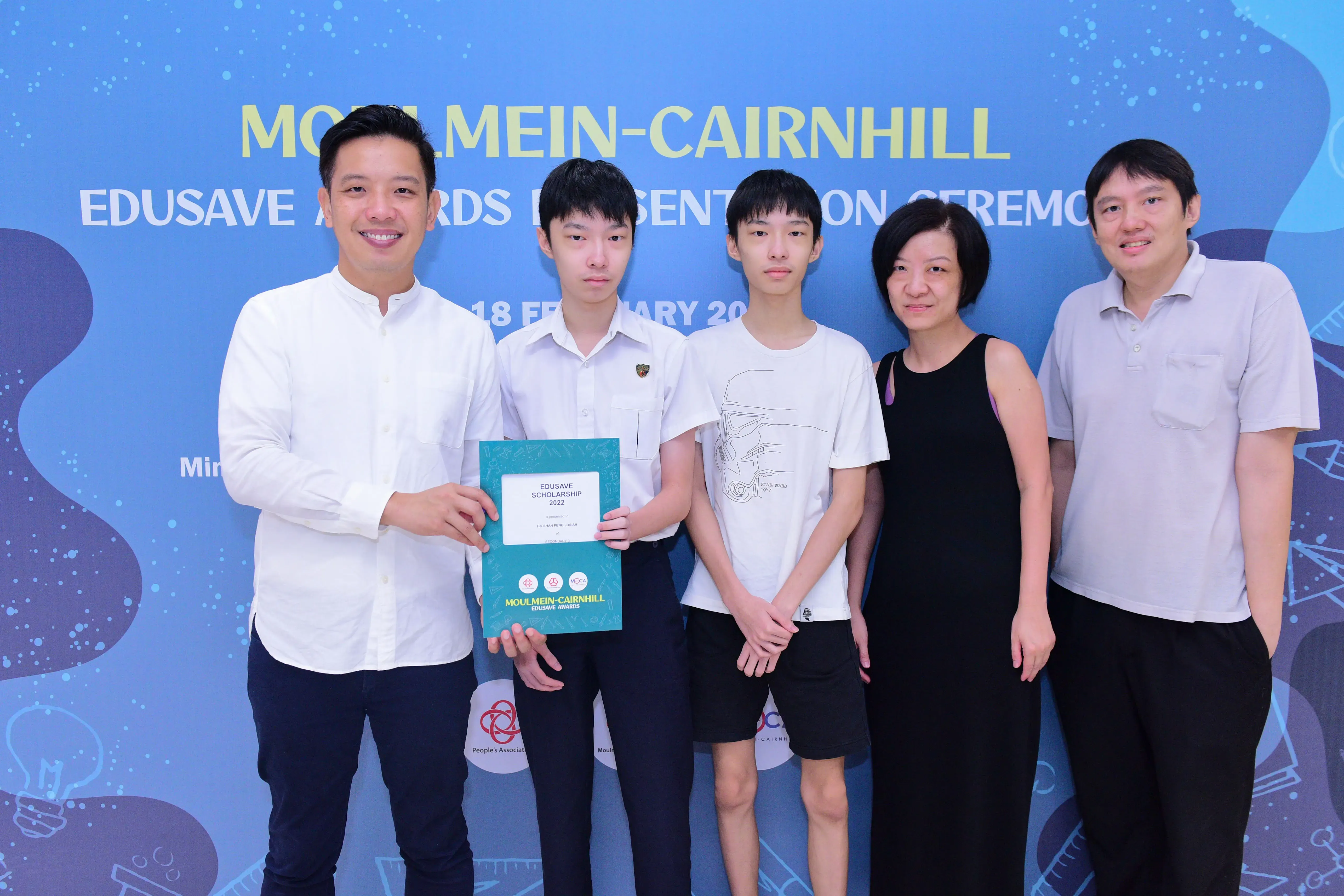 Five people pose in front of a blue Moulmein-Cairnhill Edusave Awards Presentation Ceremony banner, one holding a certificate.