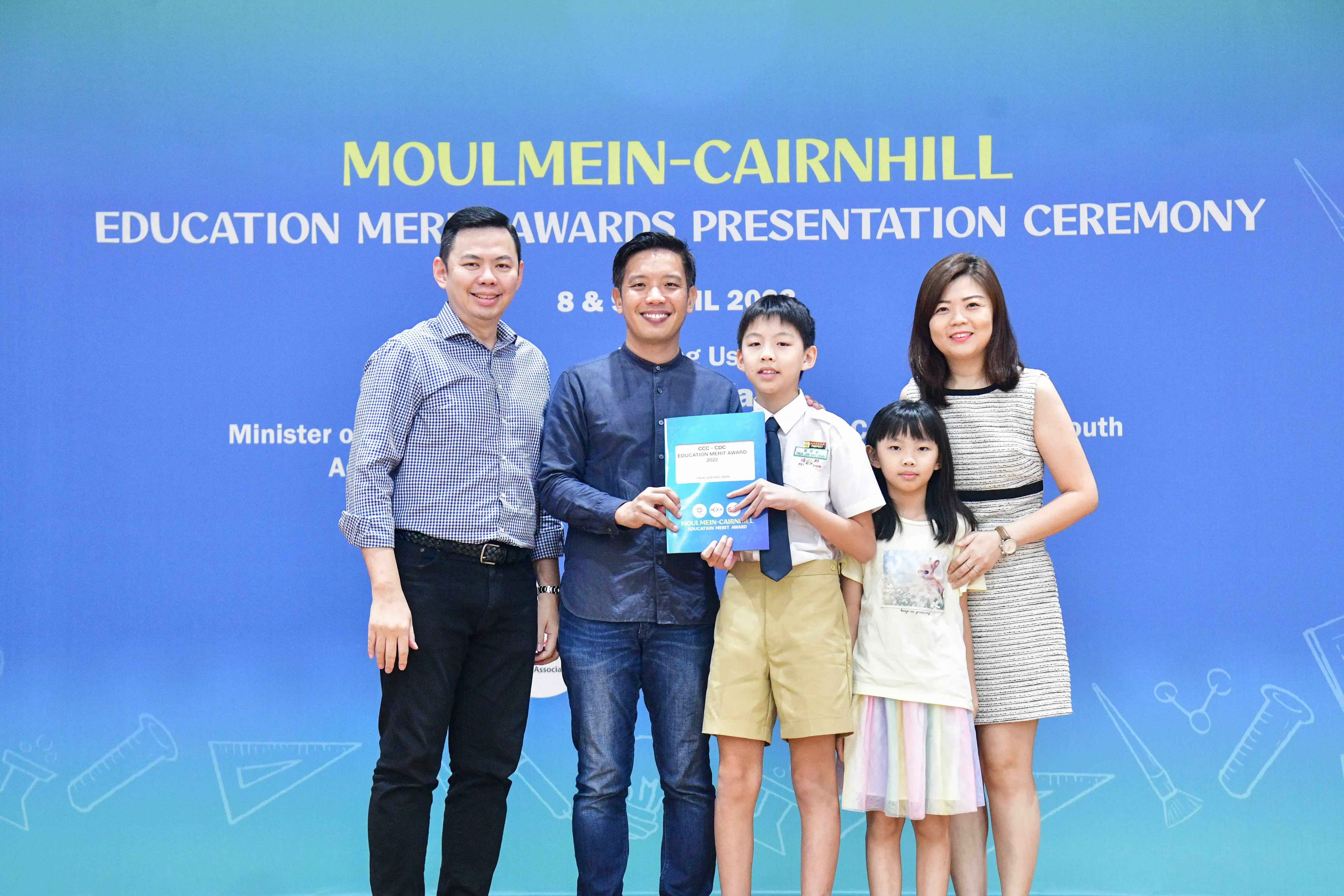 Five people pose at the Moulmein-Cairnhill Education Merit Awards Presentation Ceremony, with a boy holding an award folder.