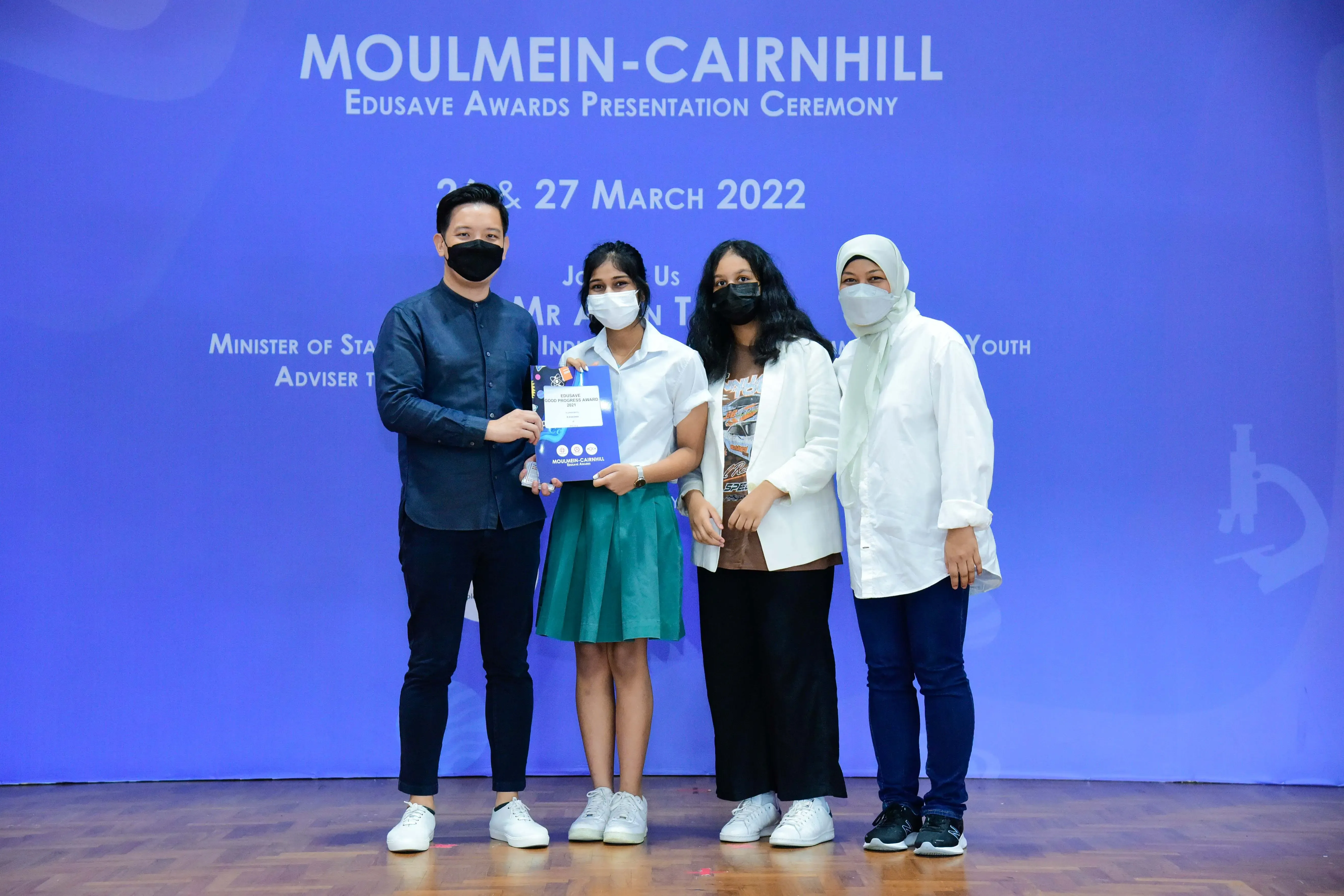 Four masked people pose on a stage at the Moulmein-Cairnhill Edusave Awards Presentation Ceremony, March 2022.