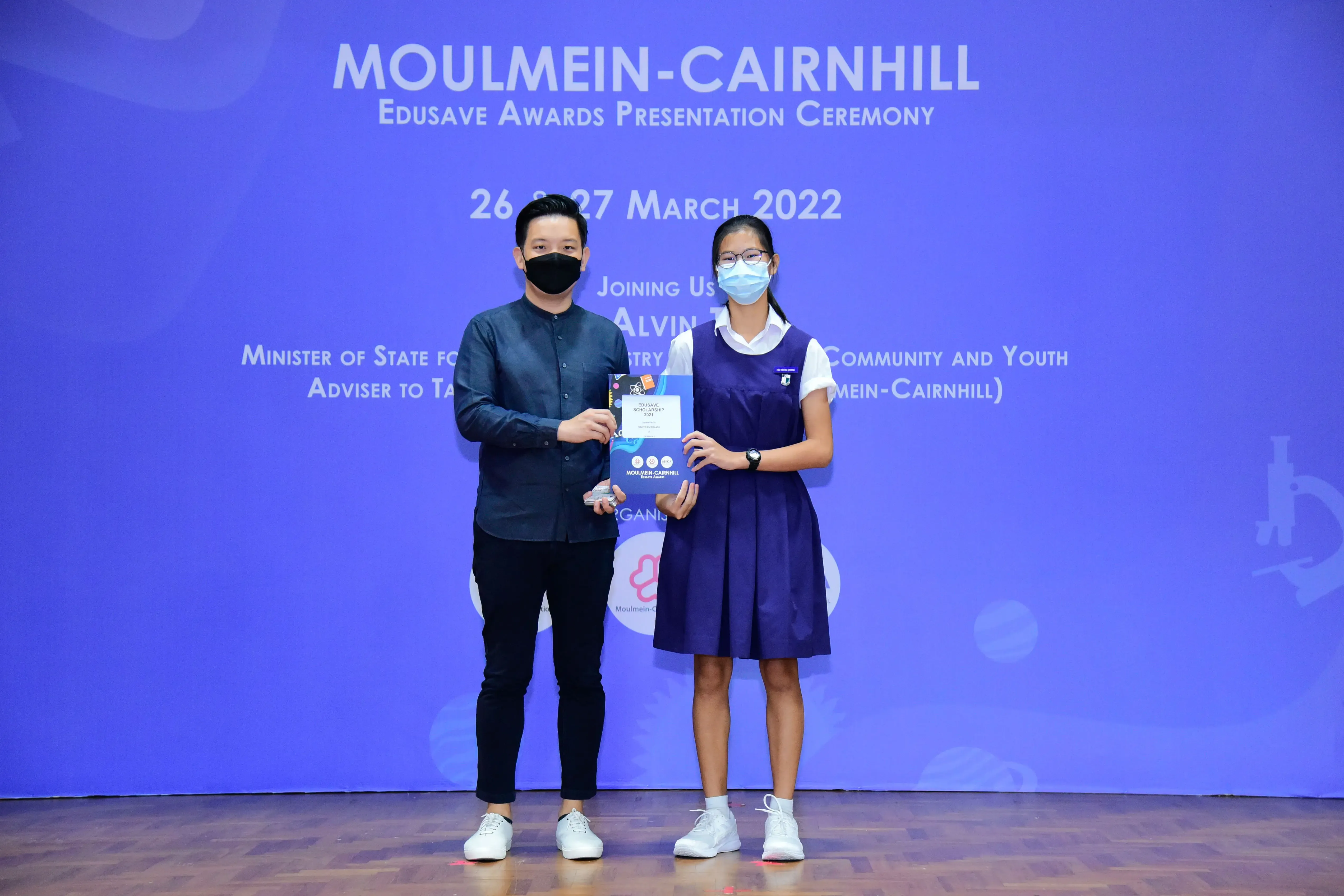 Two masked individuals hold a certificate on stage at the Moulmein-Cairnhill Edusave Awards Ceremony, March 2022.