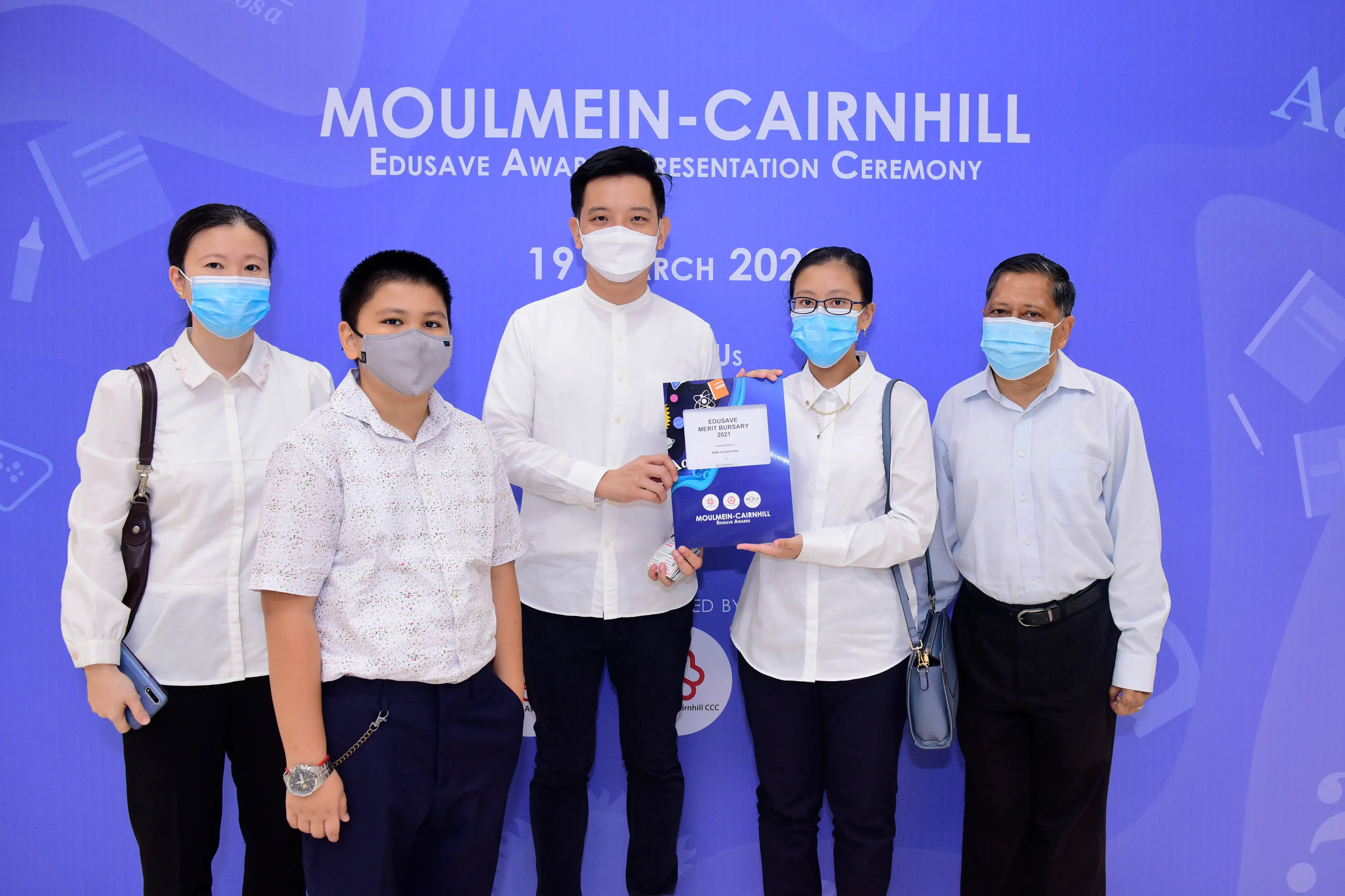 Five masked people pose at the Moulmein-Cairnhill Edusave Award Presentation Ceremony, holding an award folder.