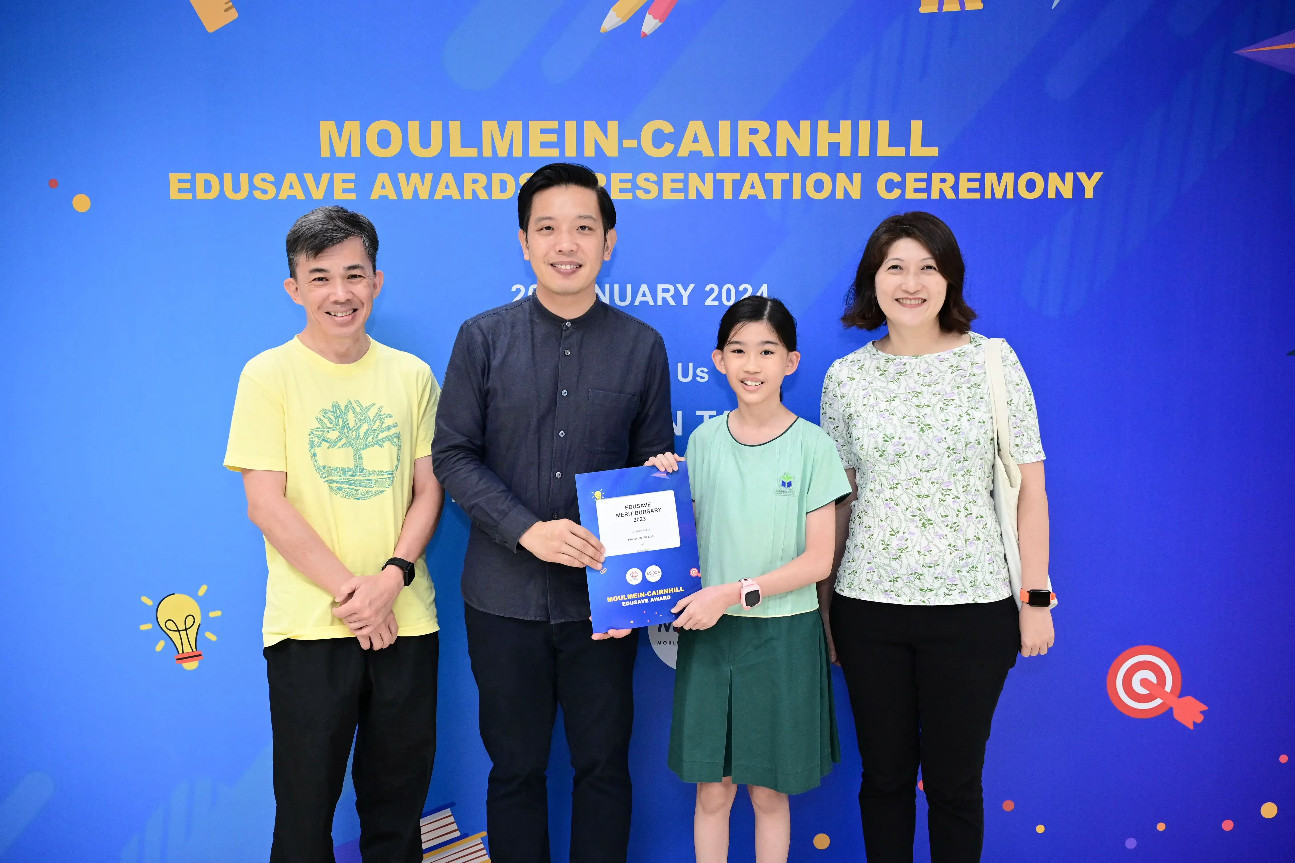 Four people pose at the Moulmein-Cairnhill Edusave Awards Ceremony, with a girl holding a blue certificate.