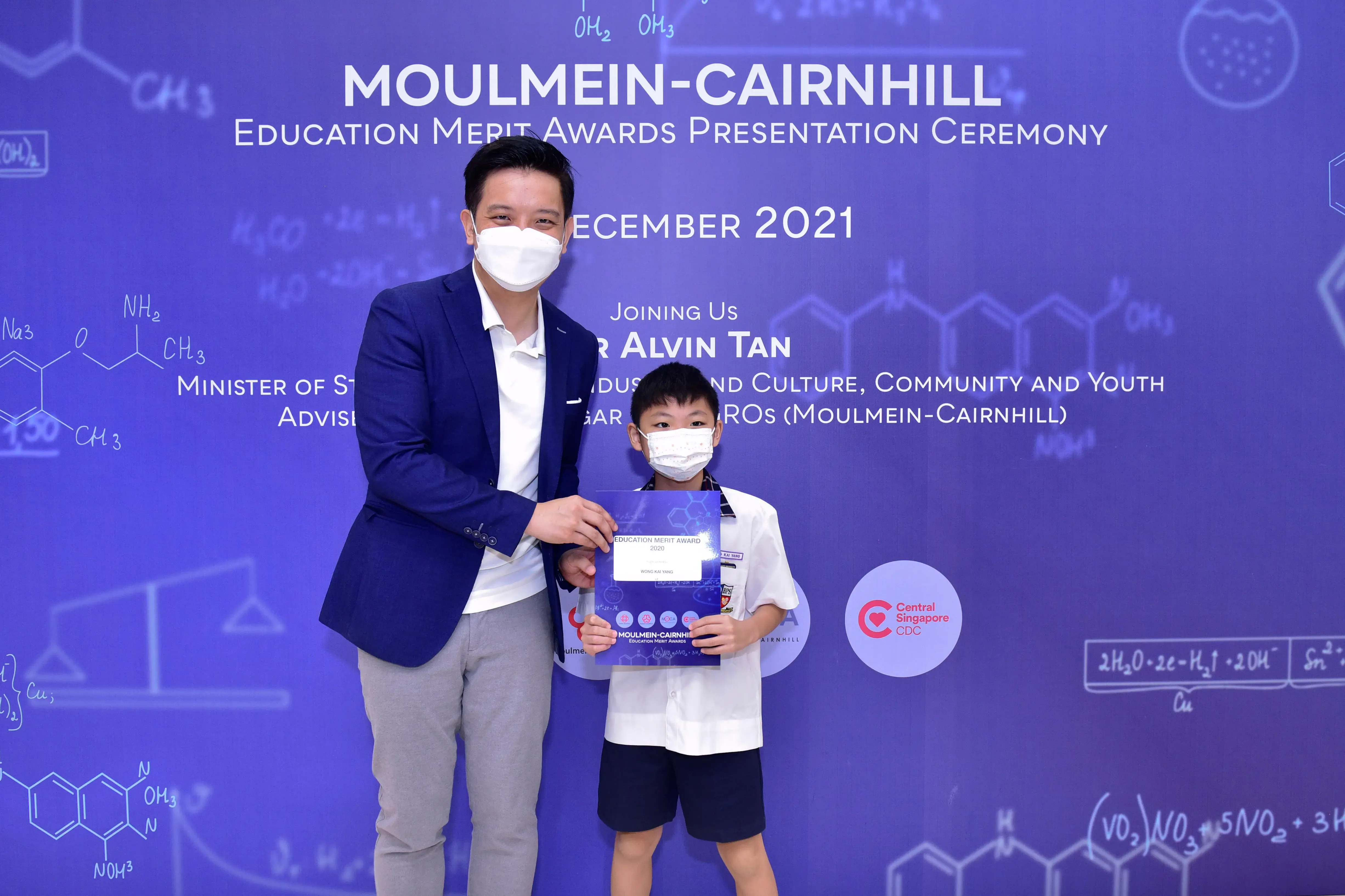 An adult and a young boy wearing masks hold a certificate at the Moulmein-Cairnhill Education Merit Awards 2021.
