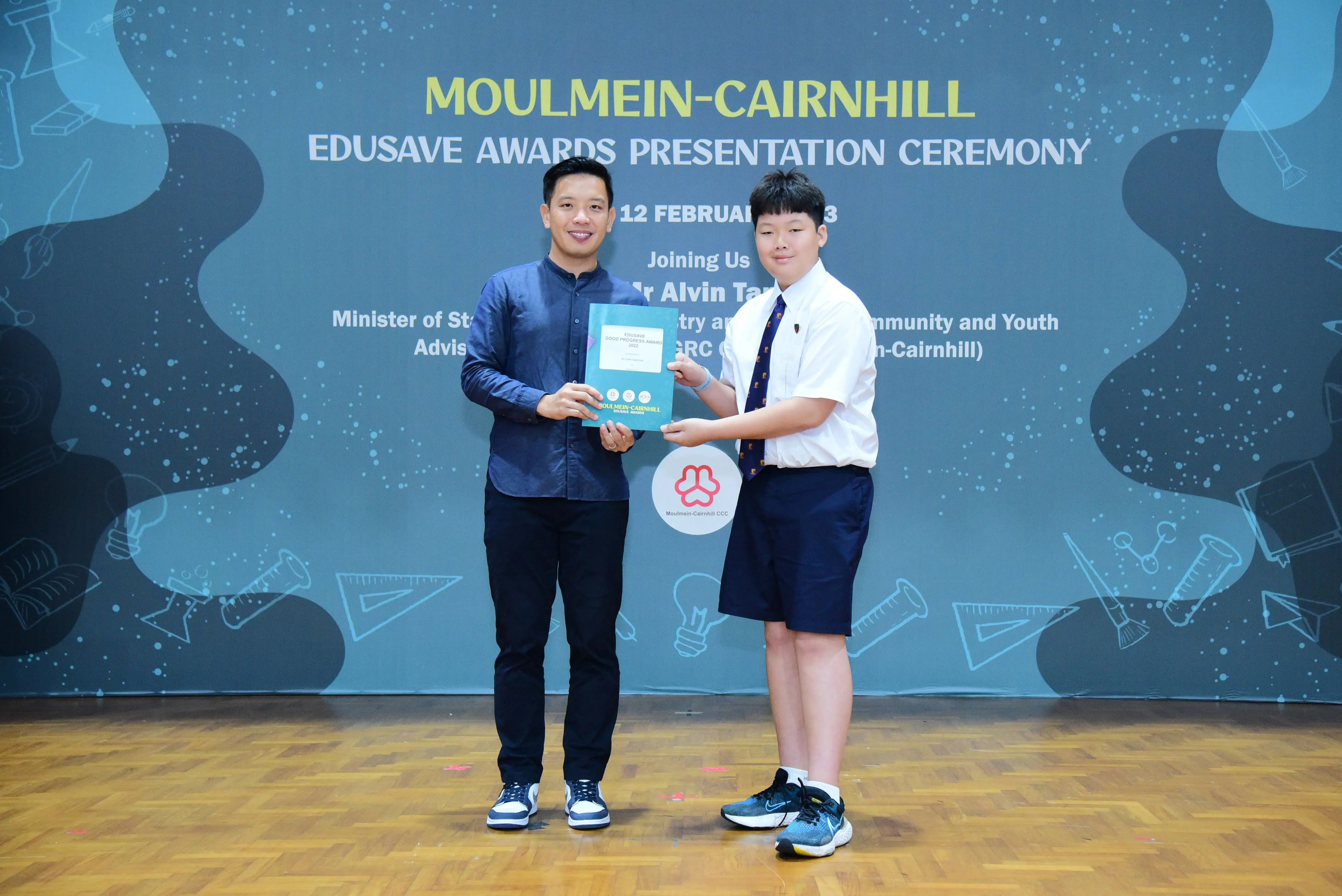 Two people hold a certificate together at the Moulmein-Cairnhill Edusave Awards Presentation Ceremony backdrop.