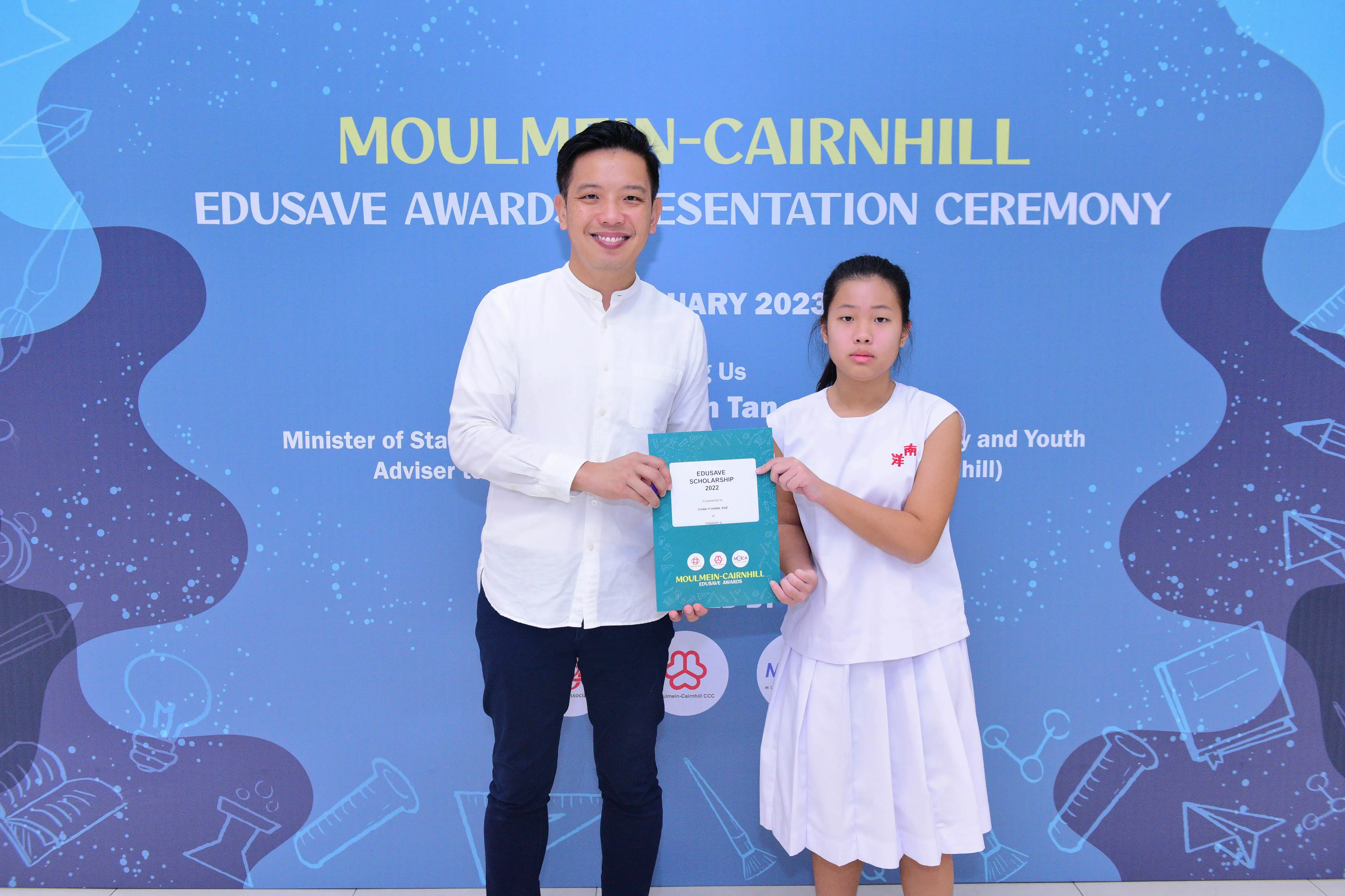 A man and a young girl hold an Edusave Scholarship certificate at the Moulmein-Cairnhill Awards Ceremony in 2023.