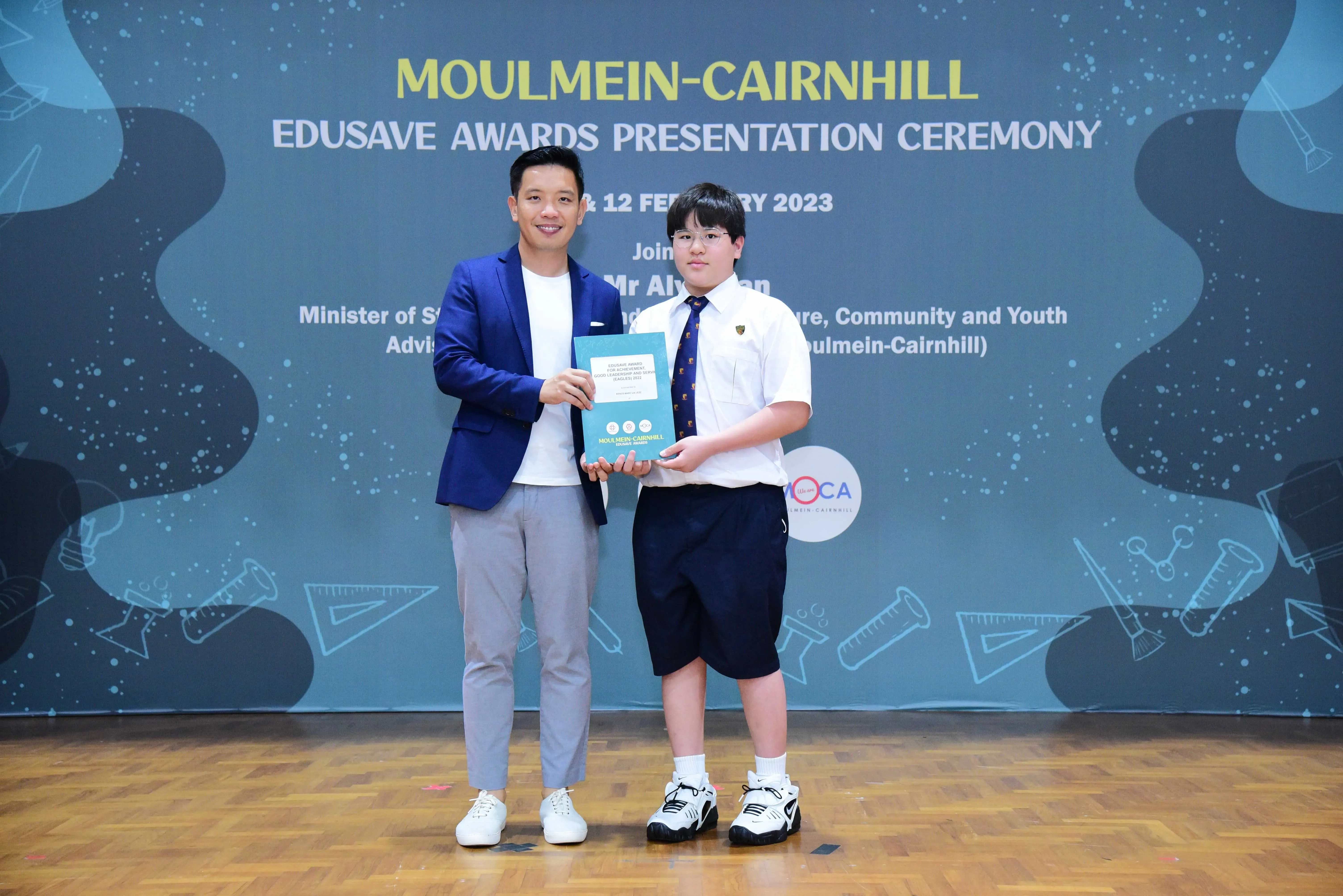 A man in a blue blazer and a uniformed boy hold a certificate at the Moulmein-Cairnhill Edusave Awards 2023.