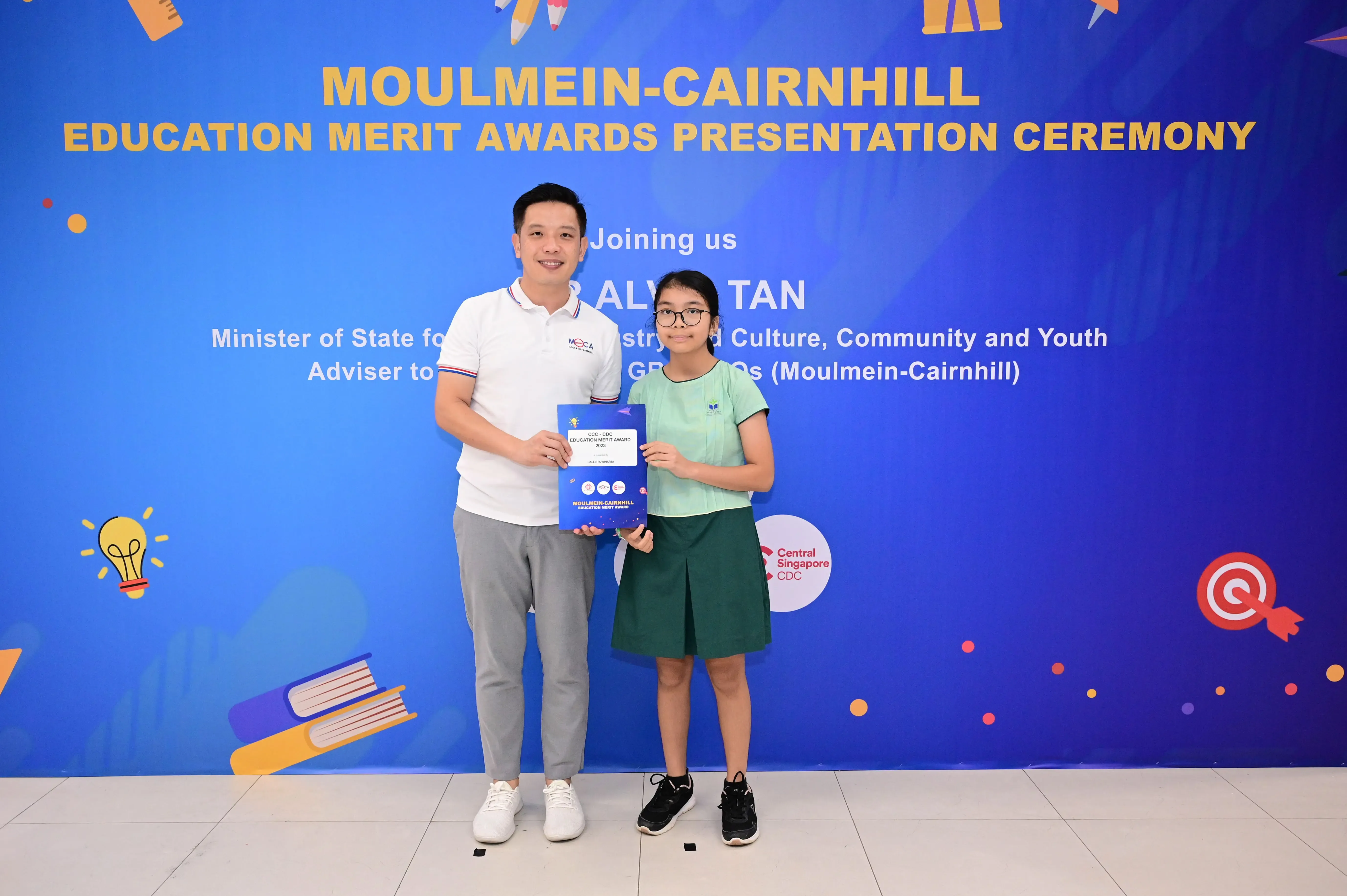 A man and a young girl hold an award certificate in front of a Moulmein-Cairnhill Education Merit Awards banner.