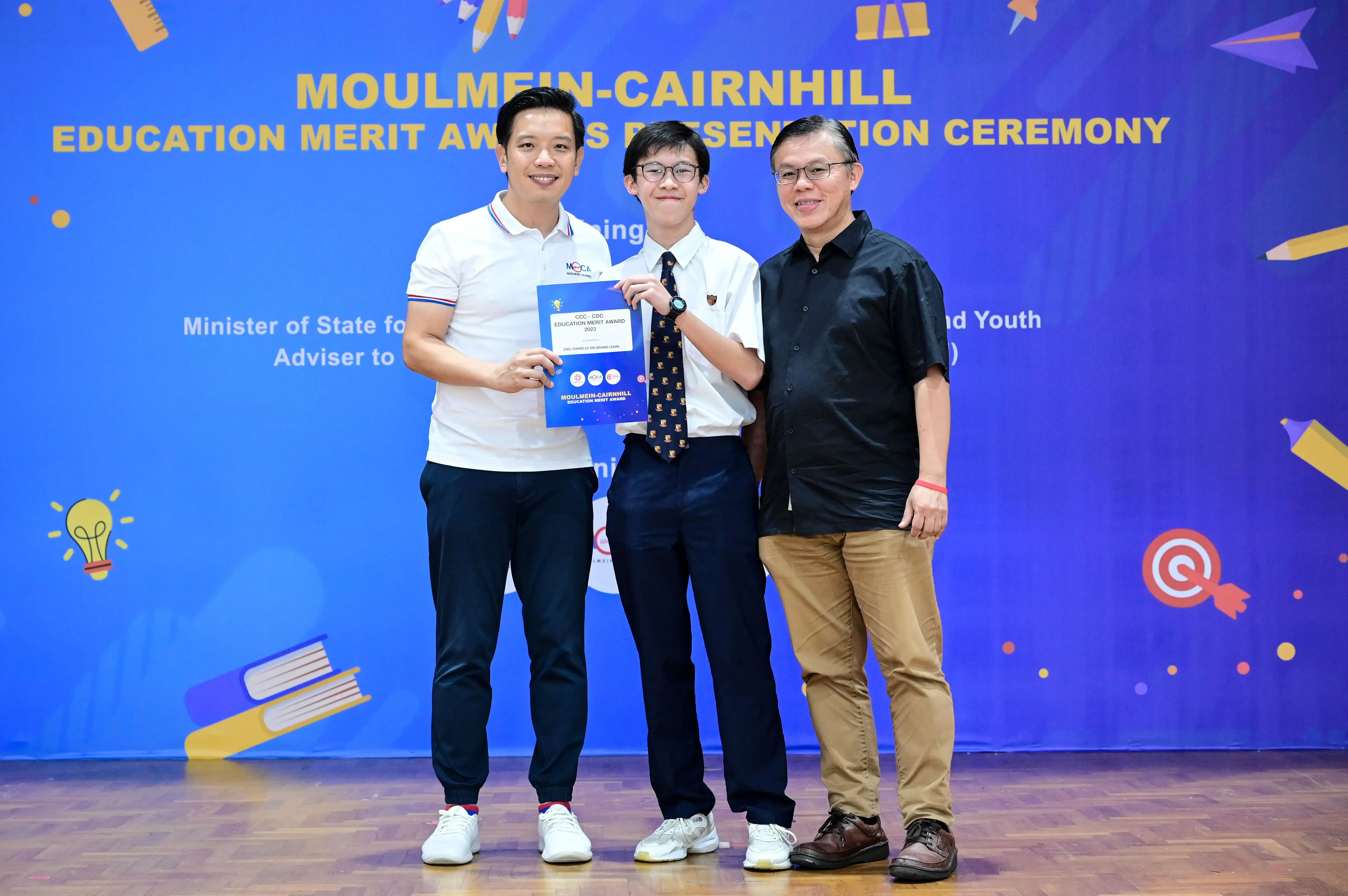 Three people stand smiling before a blue Moulmein-Cairnhill Education Merit Award Presentation Ceremony banner, holding a certificate.