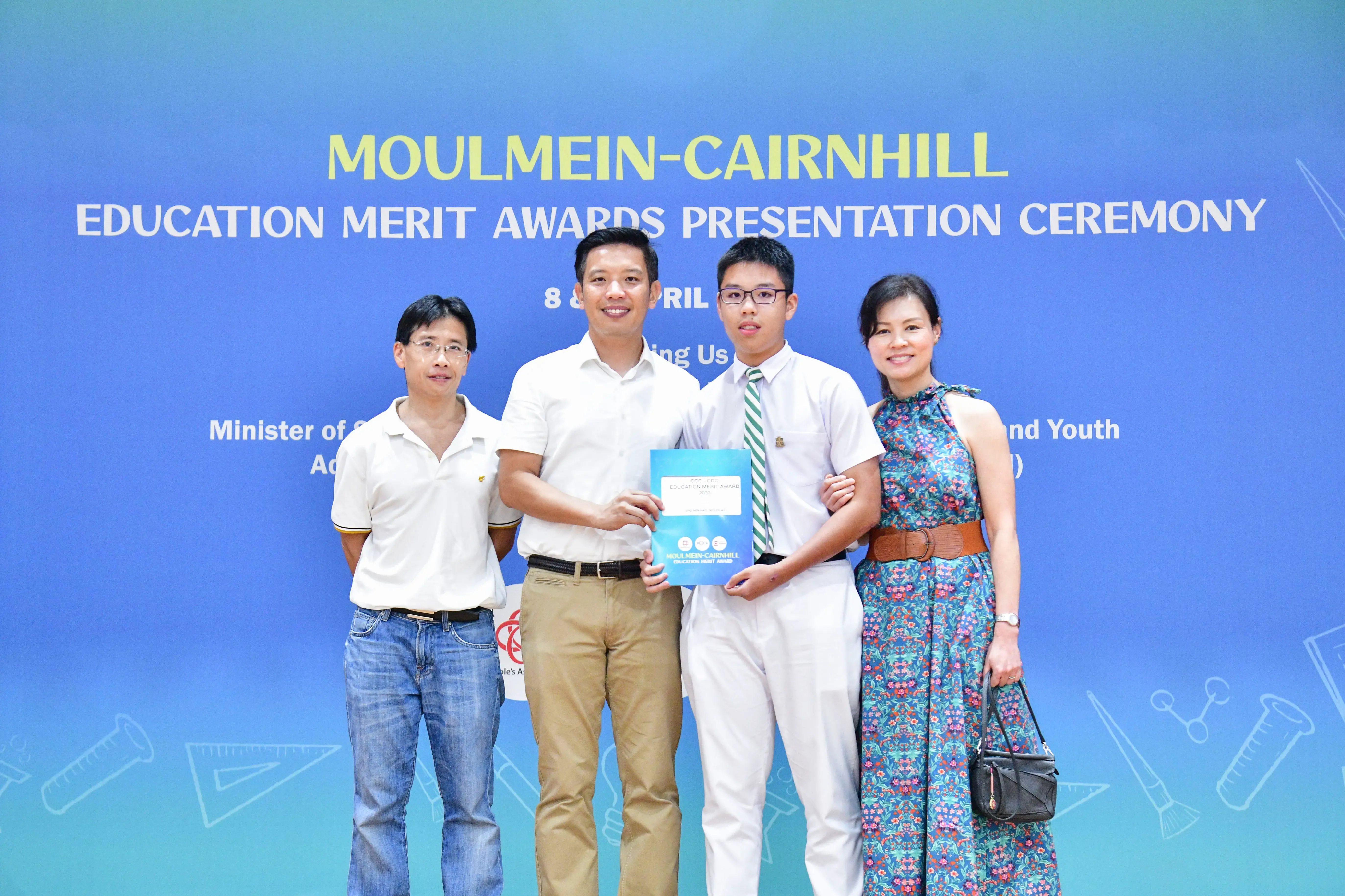 Four people pose in front of a blue Moulmein-Cairnhill Education Merit Awards Presentation Ceremony banner.