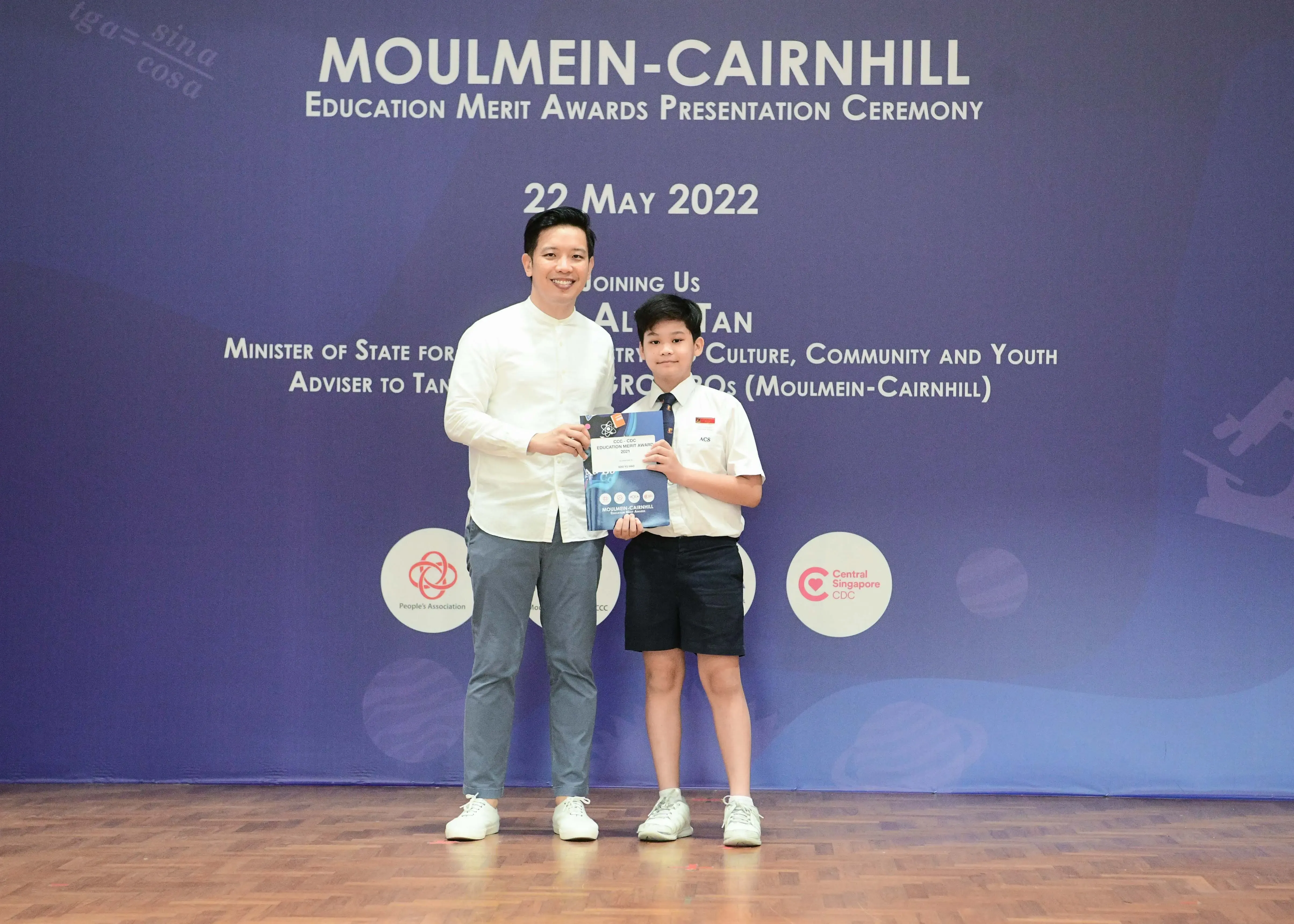 An adult and a young boy in school uniform hold an award at the Moulmein-Cairnhill Education Merit Awards Ceremony, May 2022.