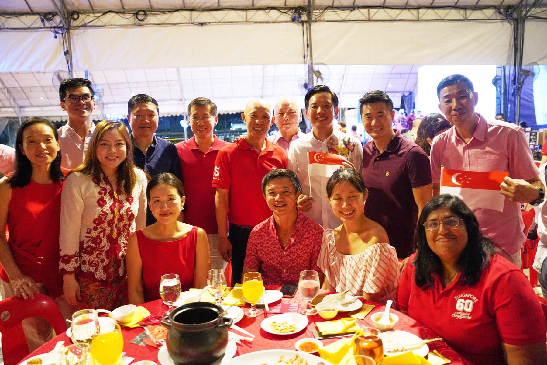 Group of people in red attire smiling at a dinner table under a tent, holding small Singapore flags.