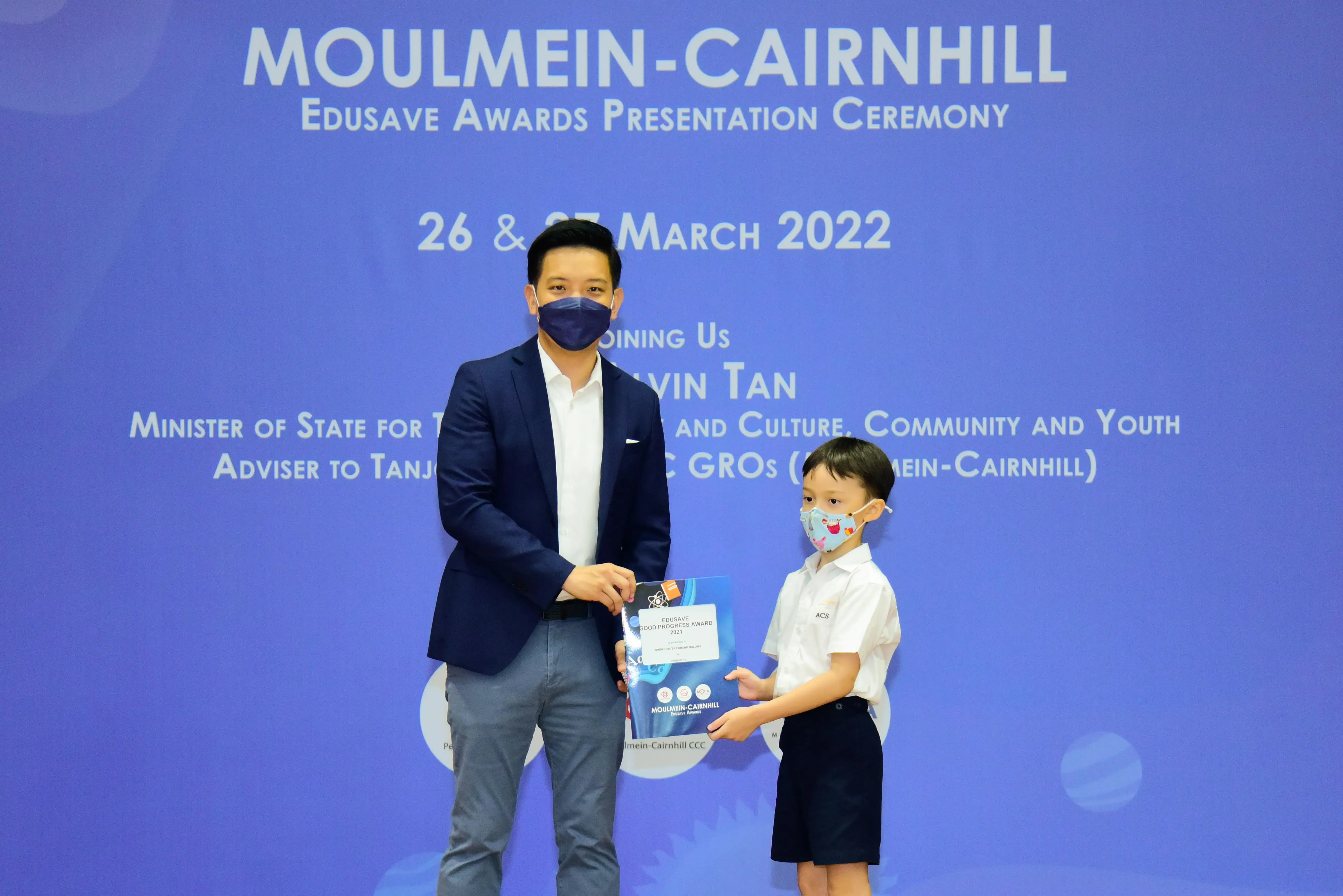 An adult and a young student in masks hold an award certificate at the Moulmein-Cairnhill Edusave Awards Ceremony 2022.