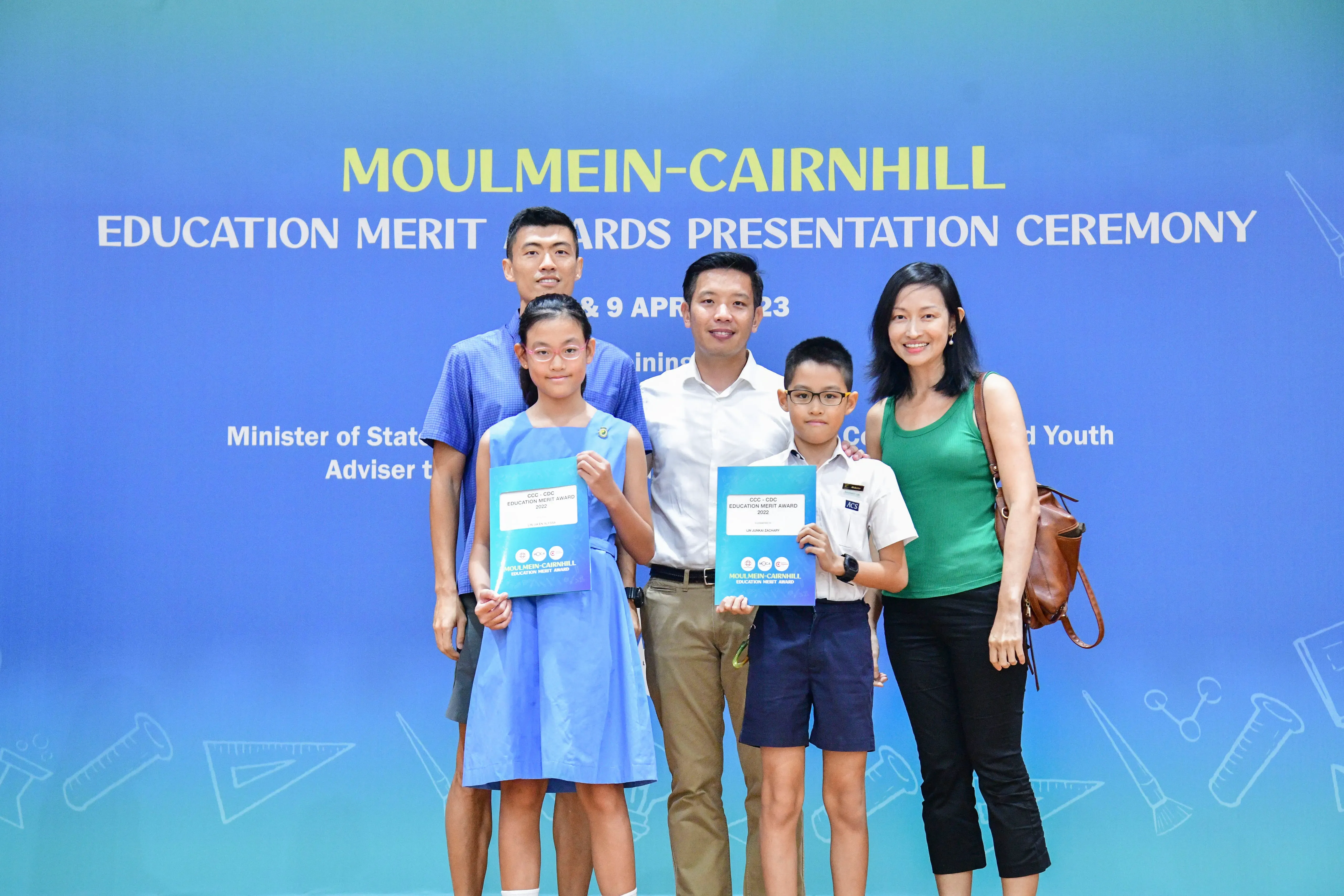 Five people pose in front of a blue Moulmein-Cairnhill Education Merit Awards banner, two children holding award folders.