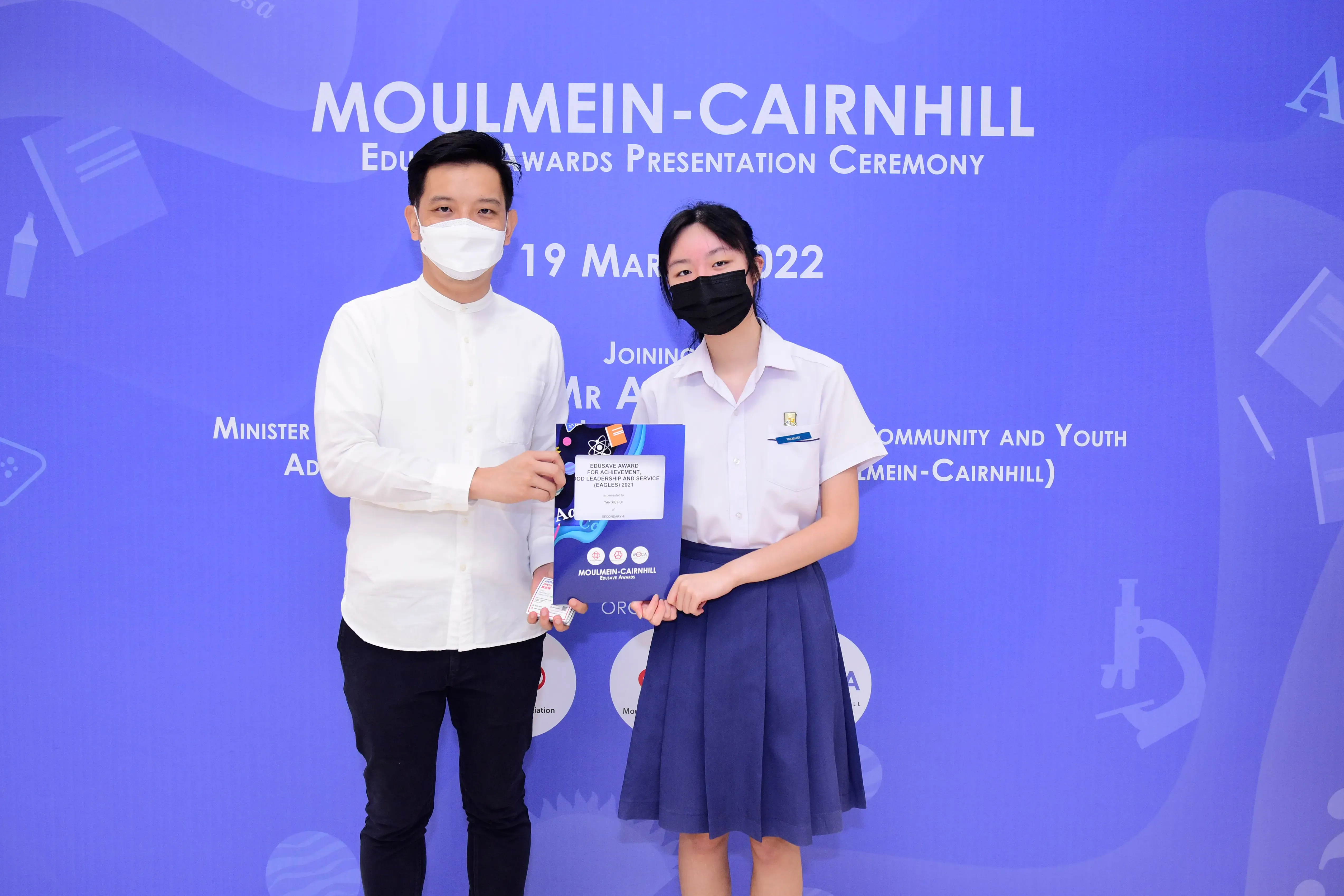 Two masked people hold an award folder at the Moulmein-Cairnhill Edusave Awards Presentation Ceremony, 19 March 2022.