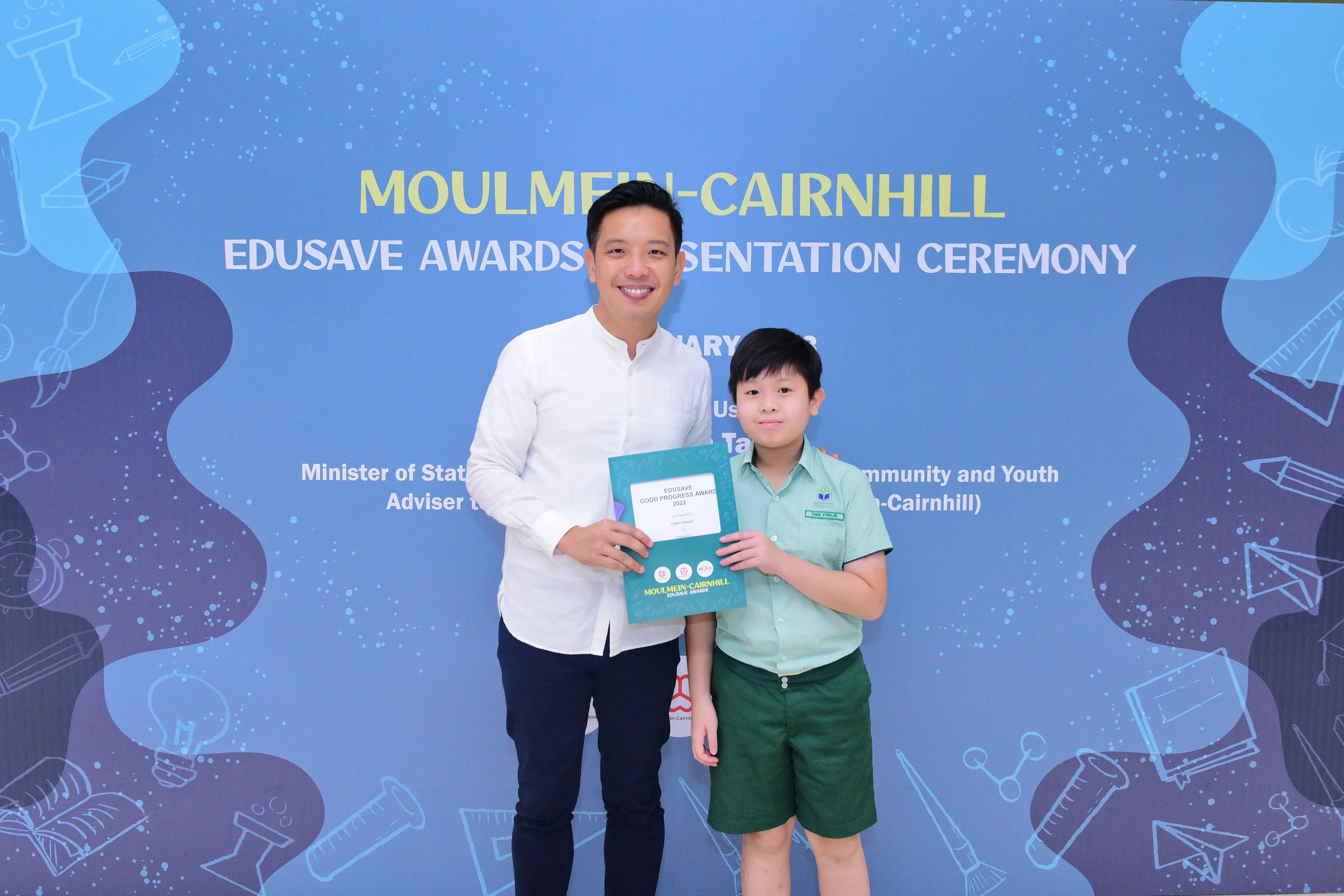Man in white shirt and young boy in green uniform hold an award certificate at Moulmein-Cairnhill Edusave ceremony.