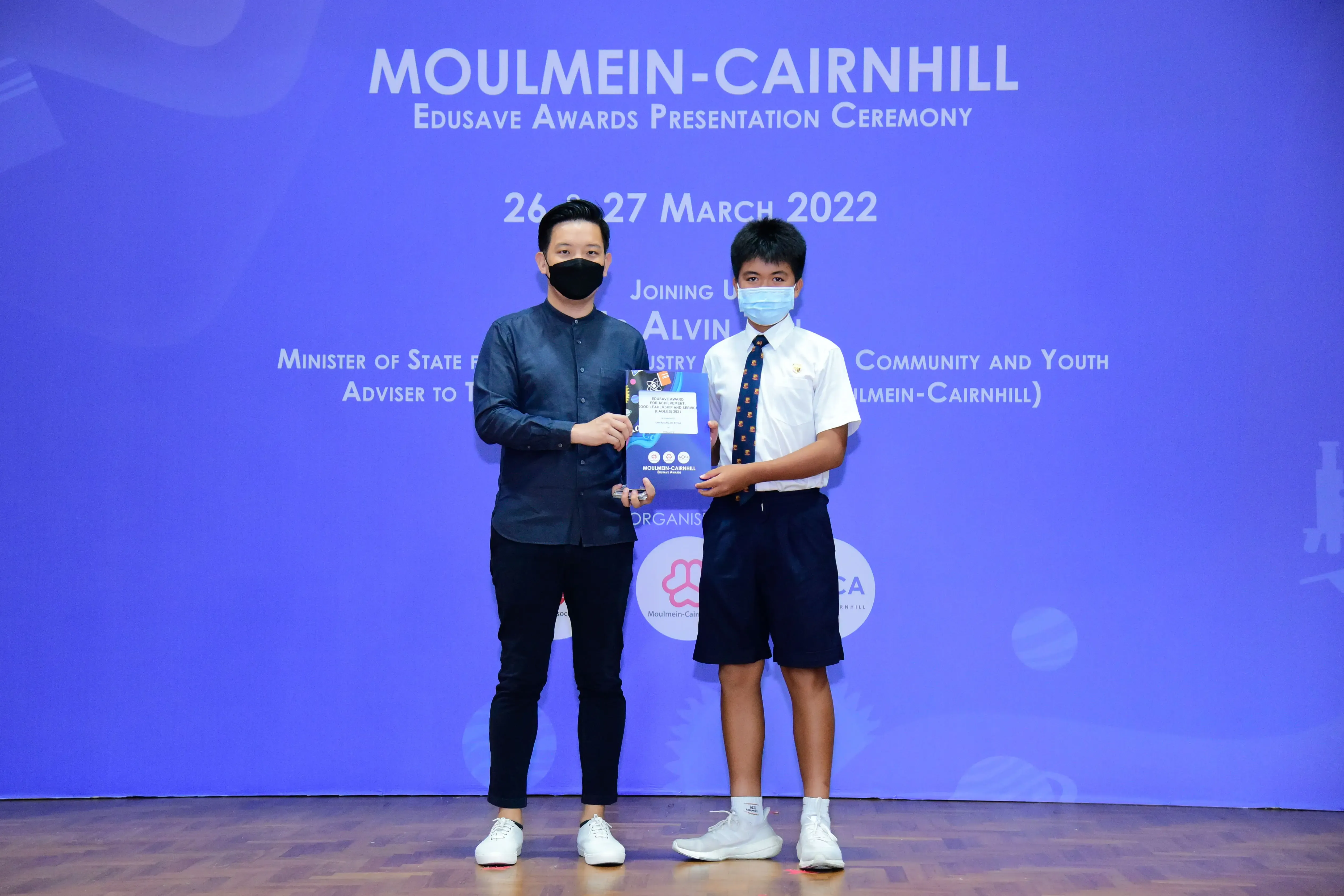 Two masked individuals hold an award booklet at the Moulmein-Cairnhill Edusave Awards Ceremony, March 2022.