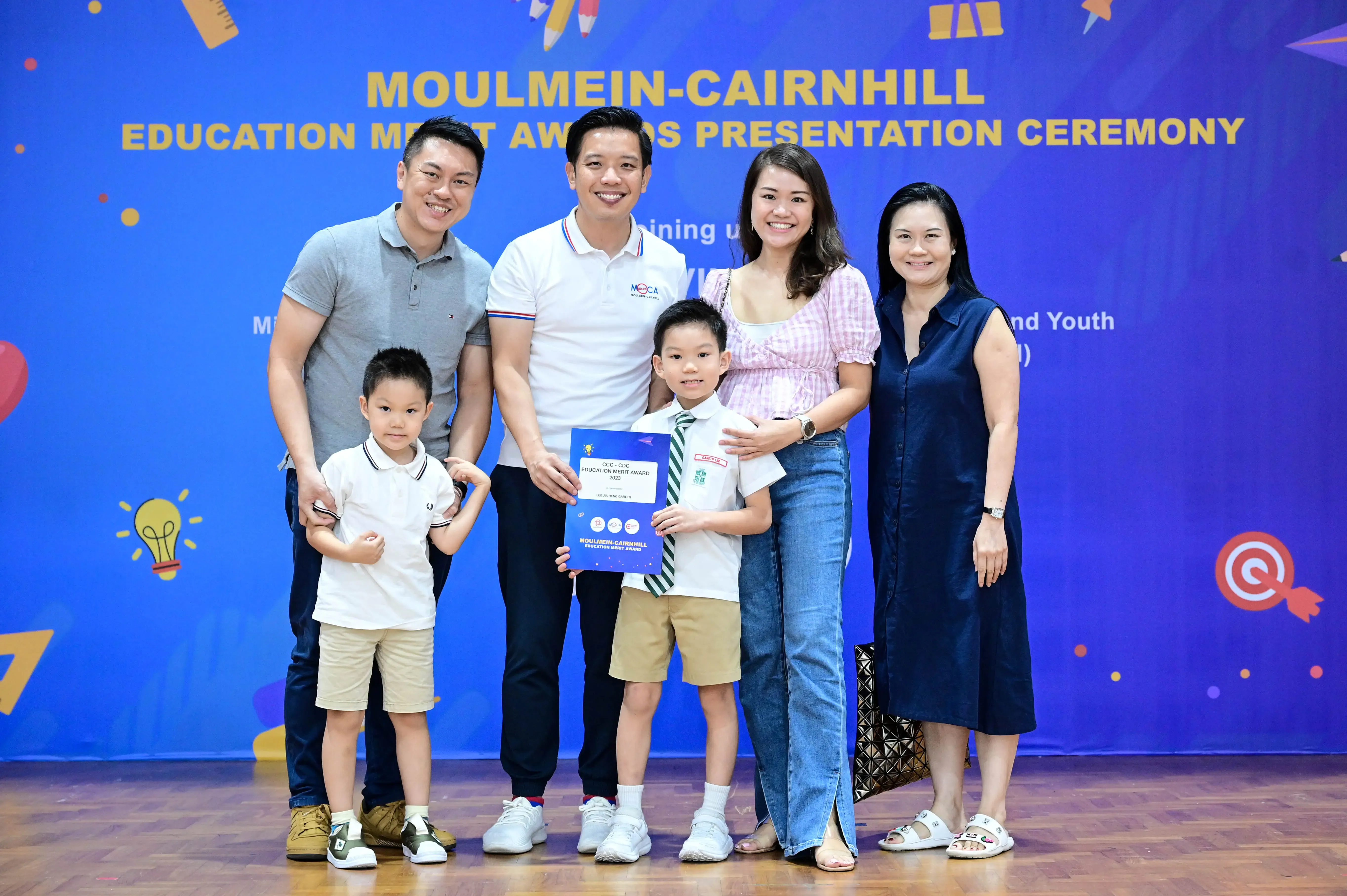Four adults and two young boys pose at the Moulmein-Cairnhill Education Merit Awards Presentation Ceremony, boy holds certificate.