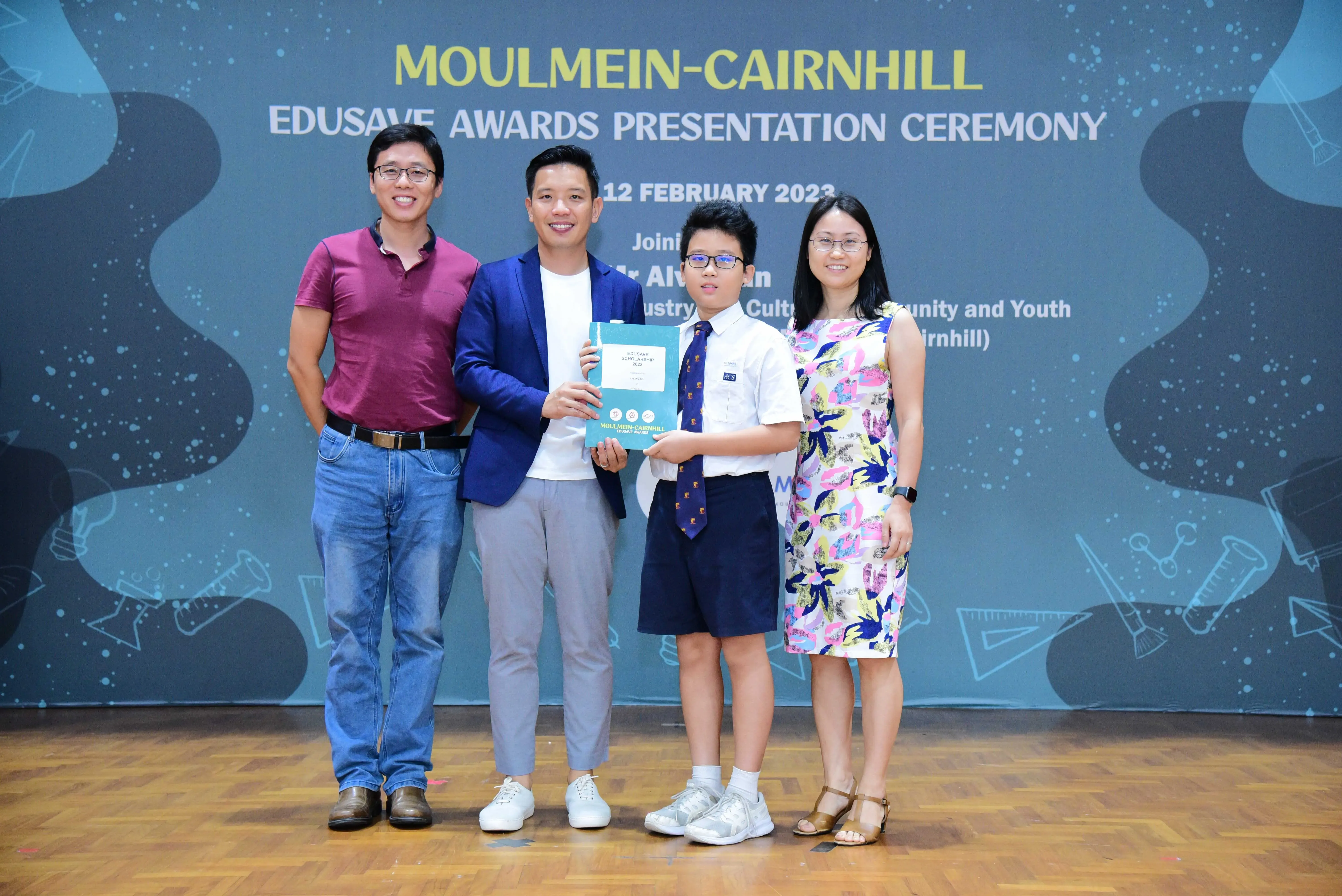 Four people pose at the Moulmein-Cairnhill Edusave Awards Presentation Ceremony, with a boy holding a certificate.