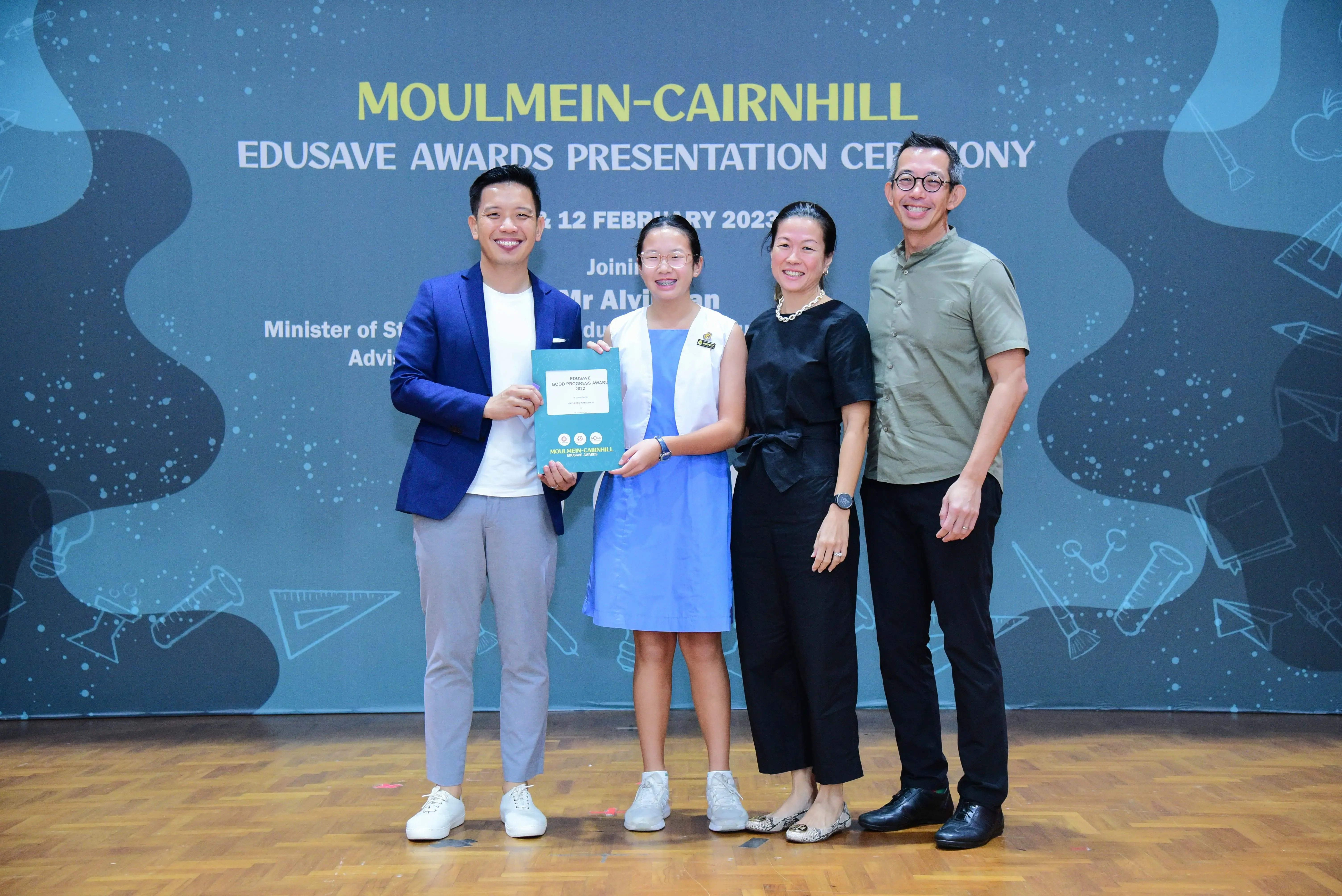 Four people pose smiling before a Moulmein-Cairnhill Edusave Awards Presentation Ceremony banner, with a girl holding an award.