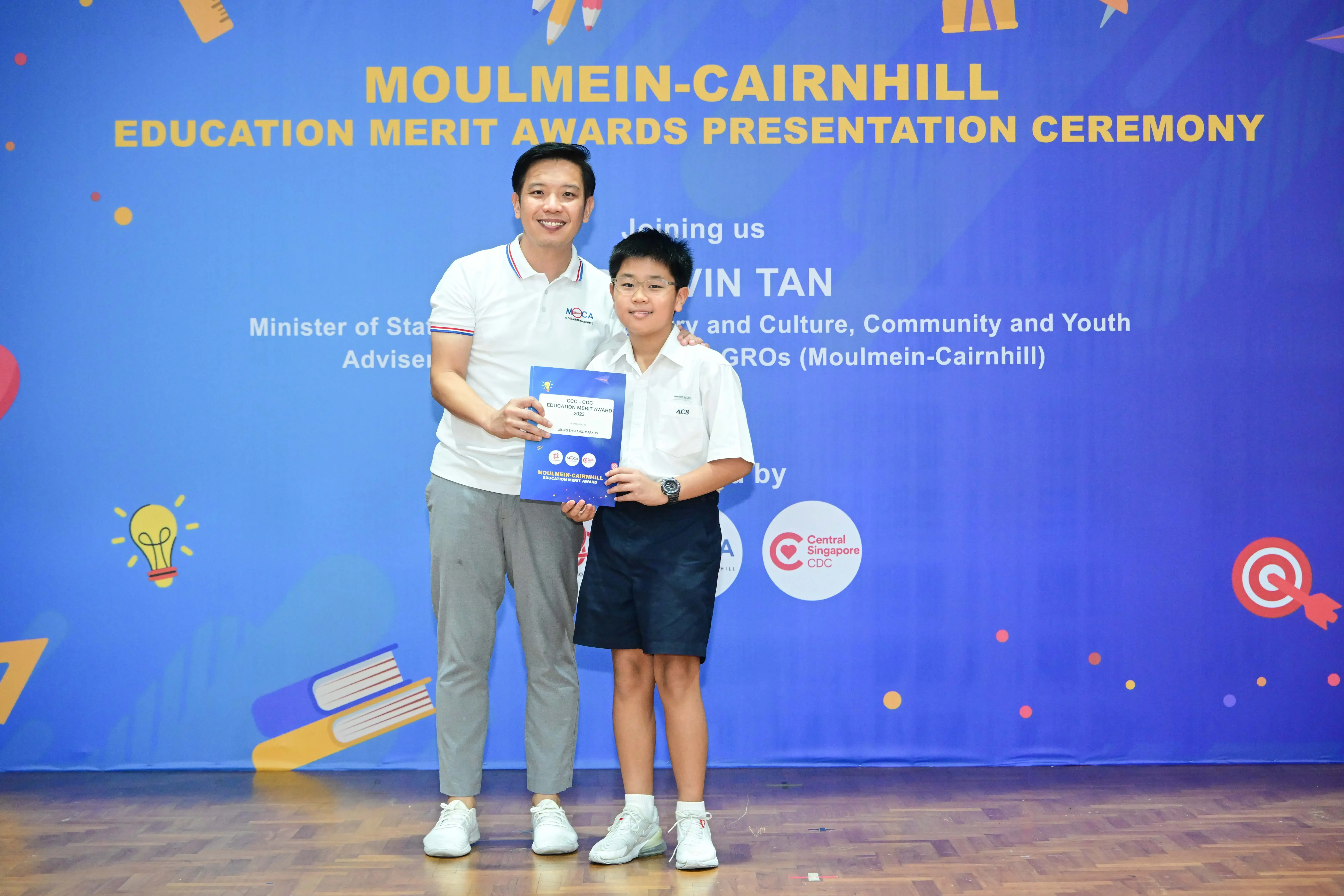 An adult and a young student pose holding a certificate at the Moulmein-Cairnhill Education Merit Awards ceremony.