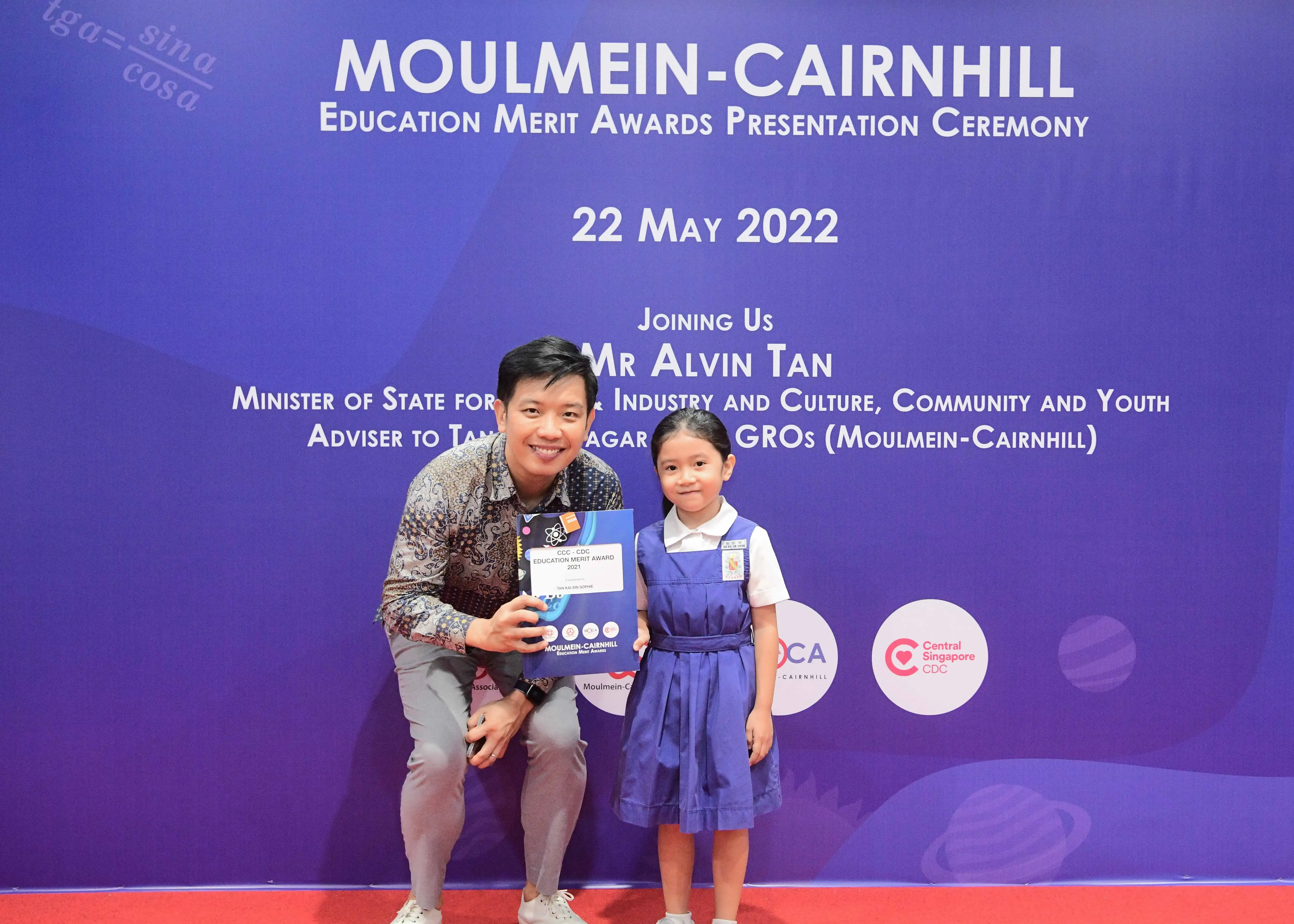 Man kneeling beside a young girl in school uniform, holding an award certificate at the Moulmein-Cairnhill Education Merit Awards 2022.