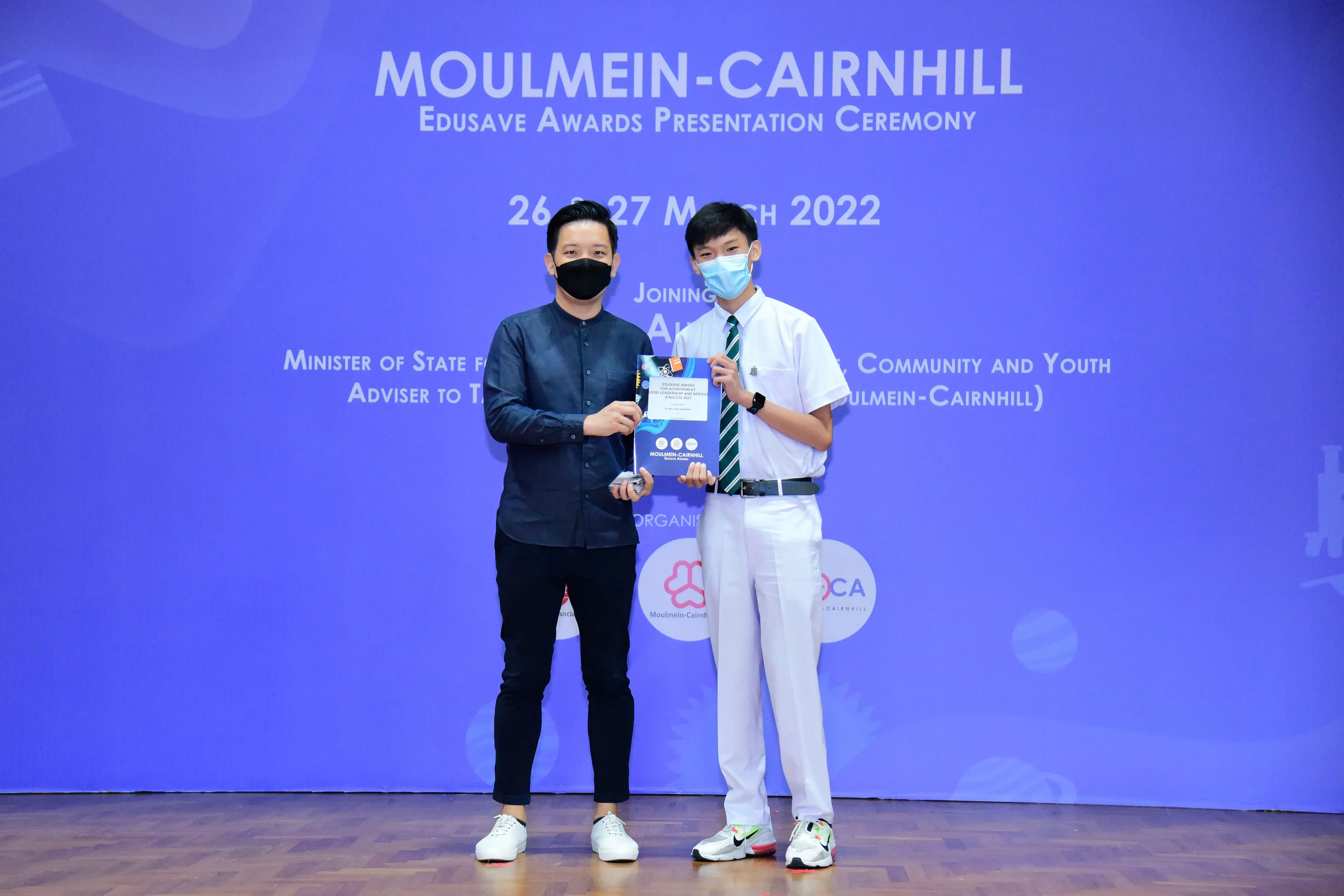Two masked men hold a certificate at the Moulmein-Cairnhill Edusave Awards Presentation Ceremony, March 2022.