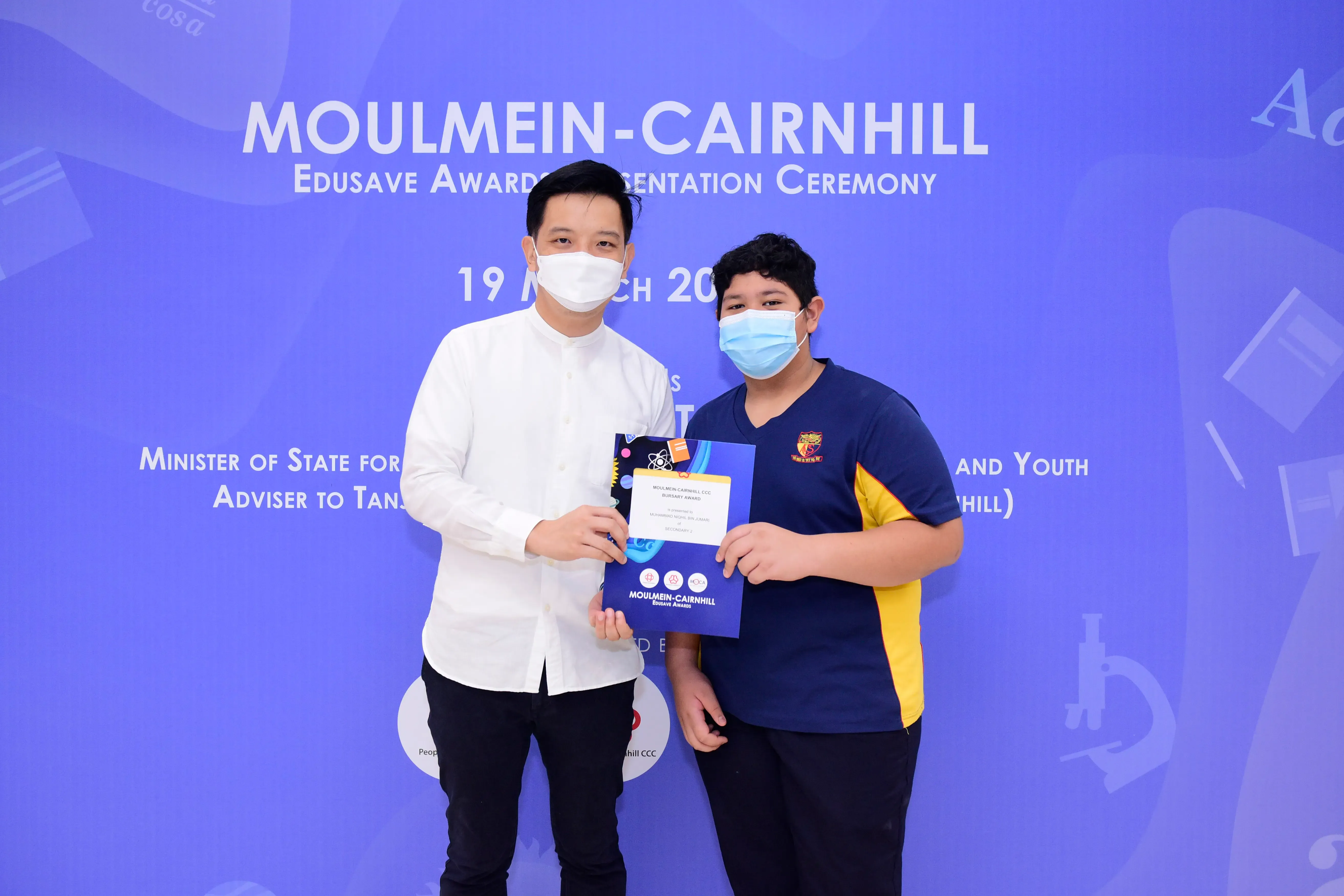 Two masked individuals hold a certificate at the Moulmein-Cairnhill Edusave Awards Presentation Ceremony.