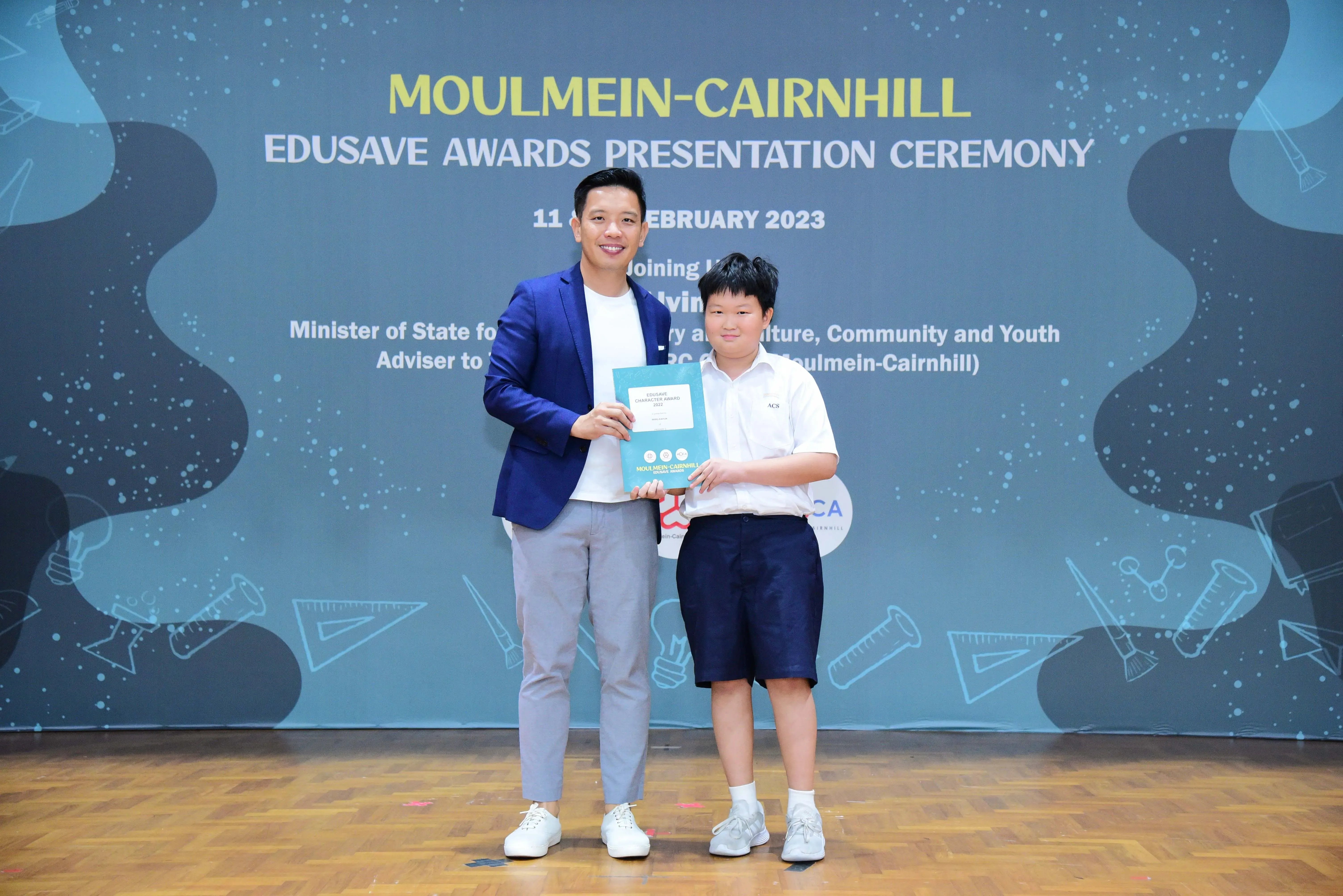 A man in a blue blazer and a young boy in school uniform hold a certificate at the Moulmein-Cairnhill Edusave Awards 2023.