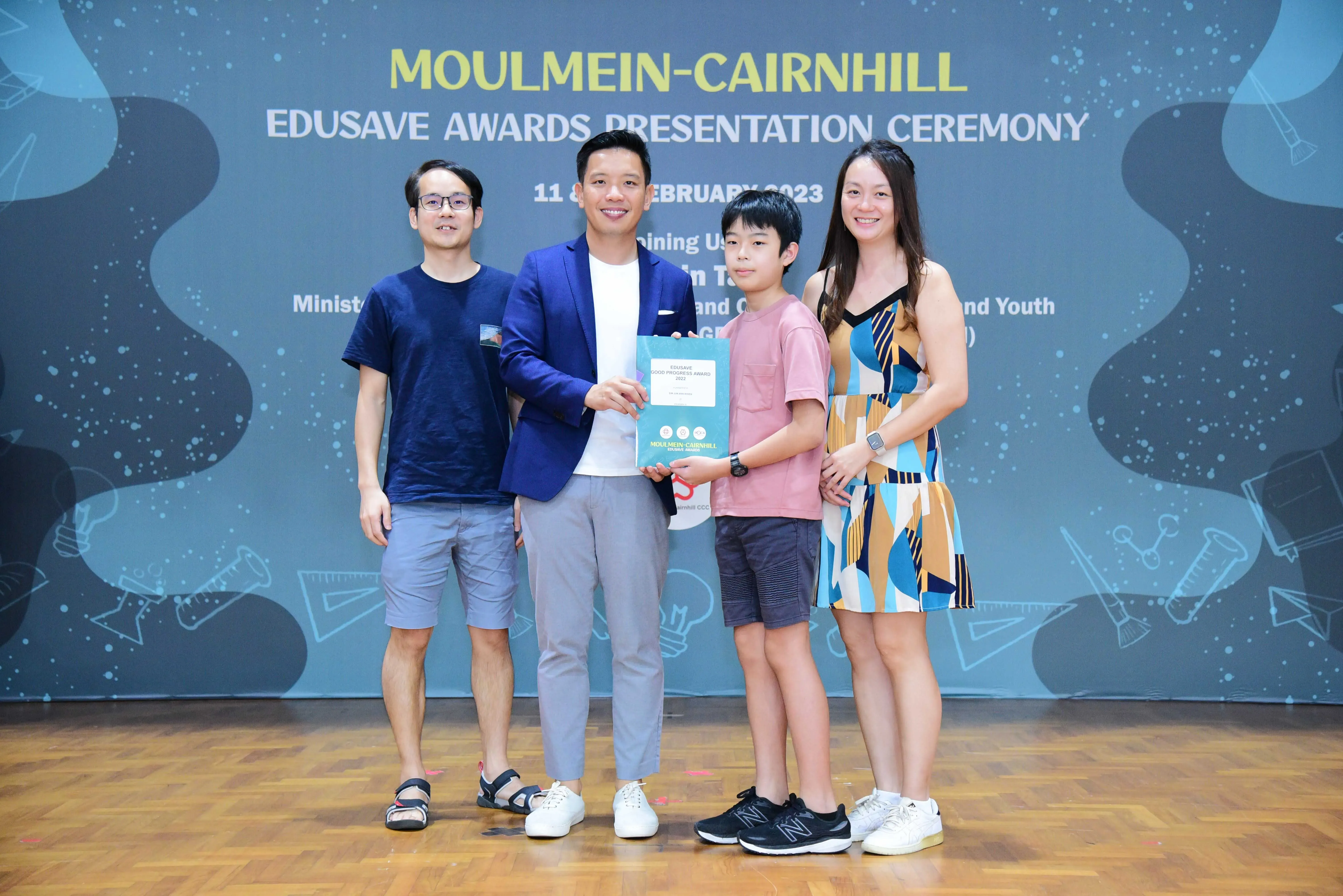 Four people pose at the Moulmein-Cairnhill Edusave Awards Presentation Ceremony, with a boy holding an award folder.