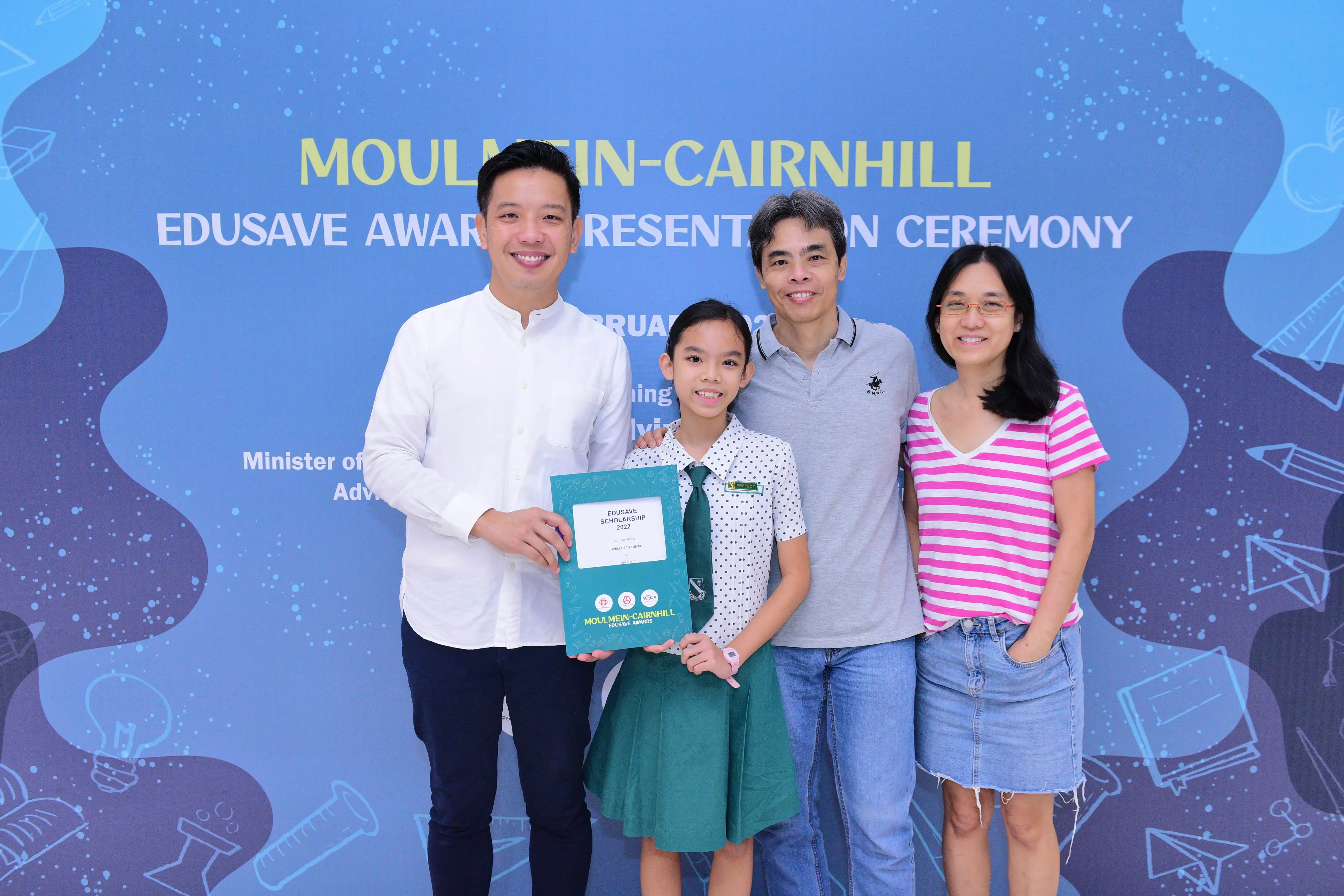 A girl in school uniform holds an Edusave Scholarship certificate, posing with three adults at the Moulmein-Cairnhill awards ceremony.