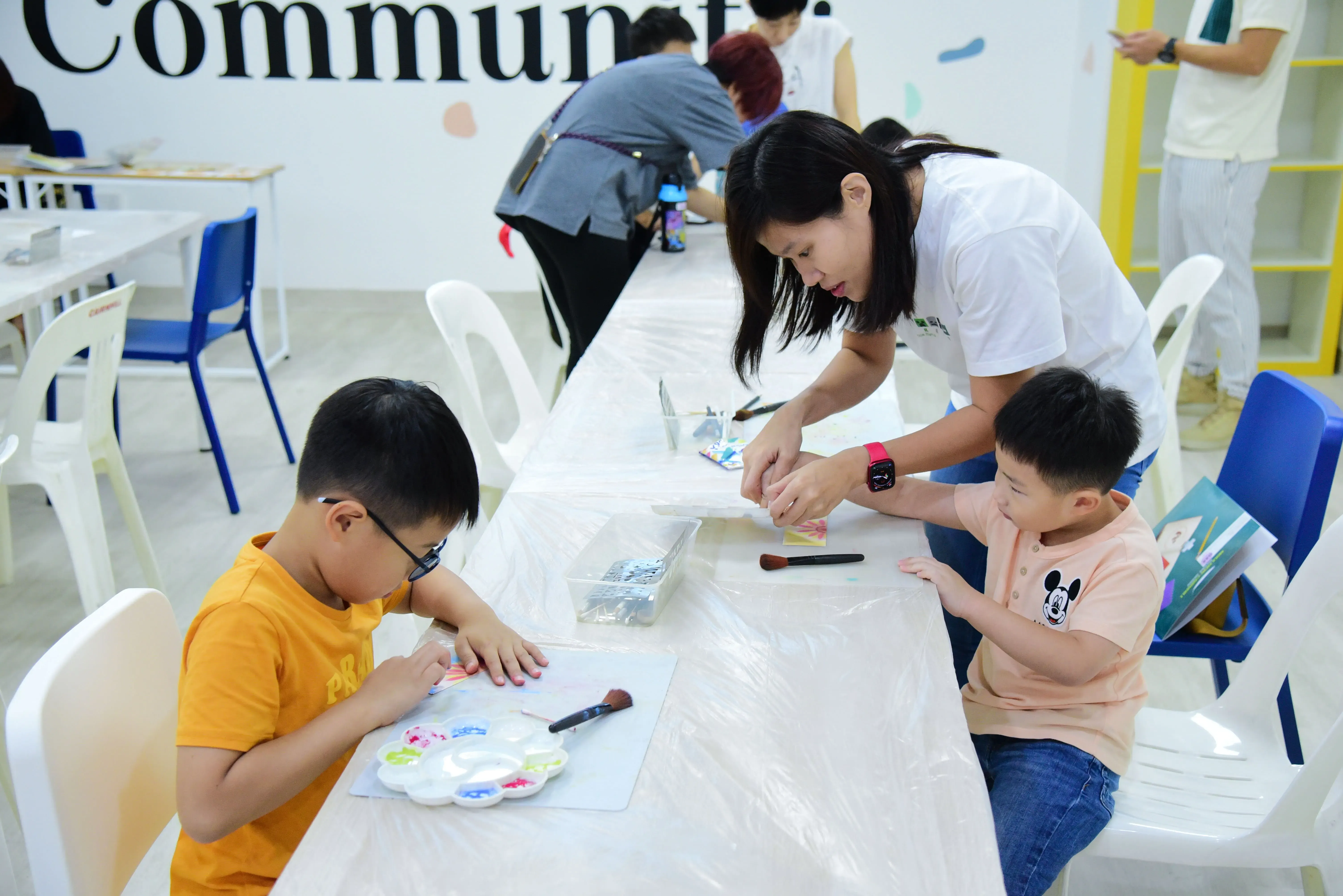 A woman helps two young boys with a painting craft activity at a table covered with plastic sheeting.