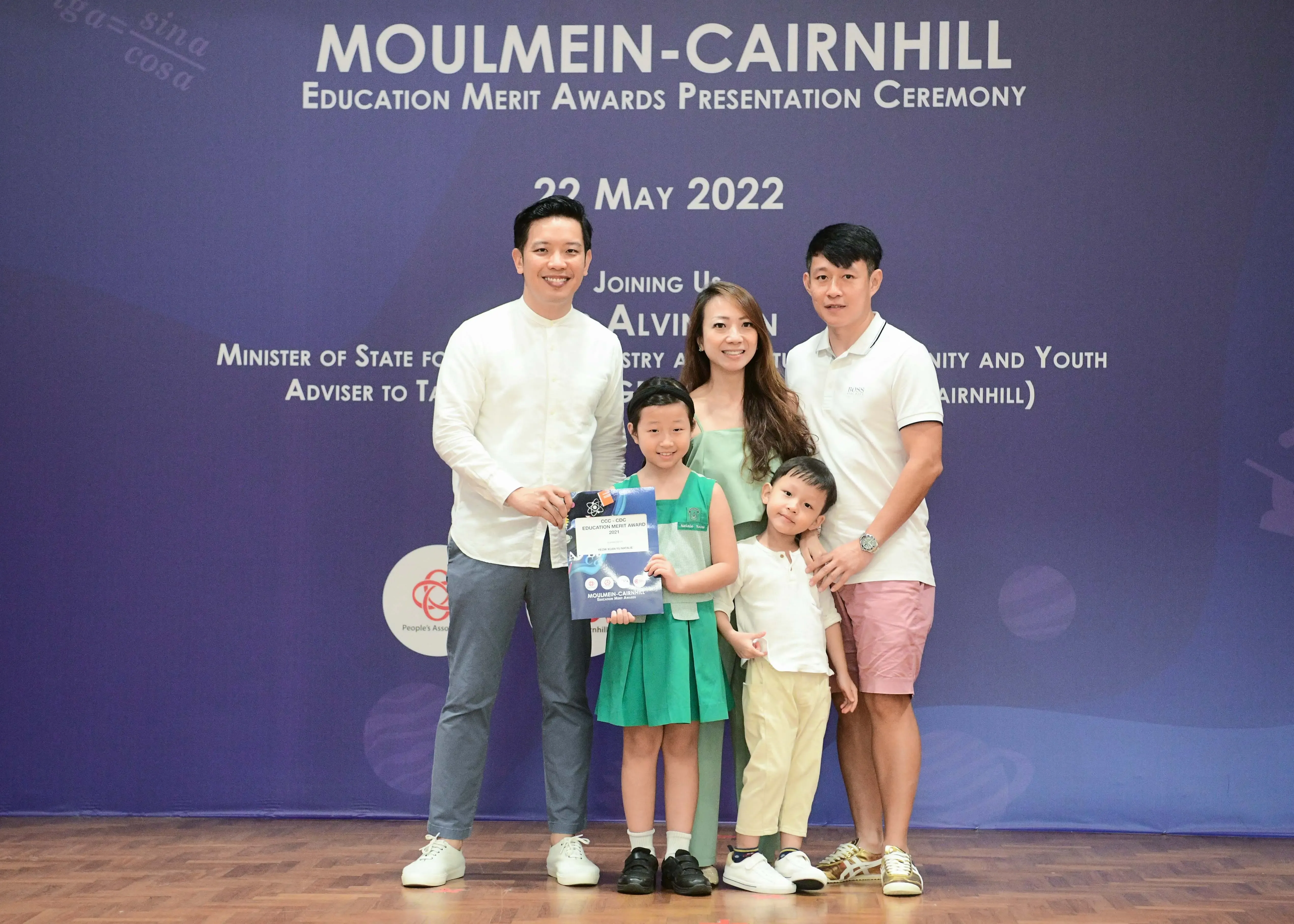 Five people, including two children, pose before a Moulmein-Cairnhill Education Merit Awards banner dated 22 May 2022.