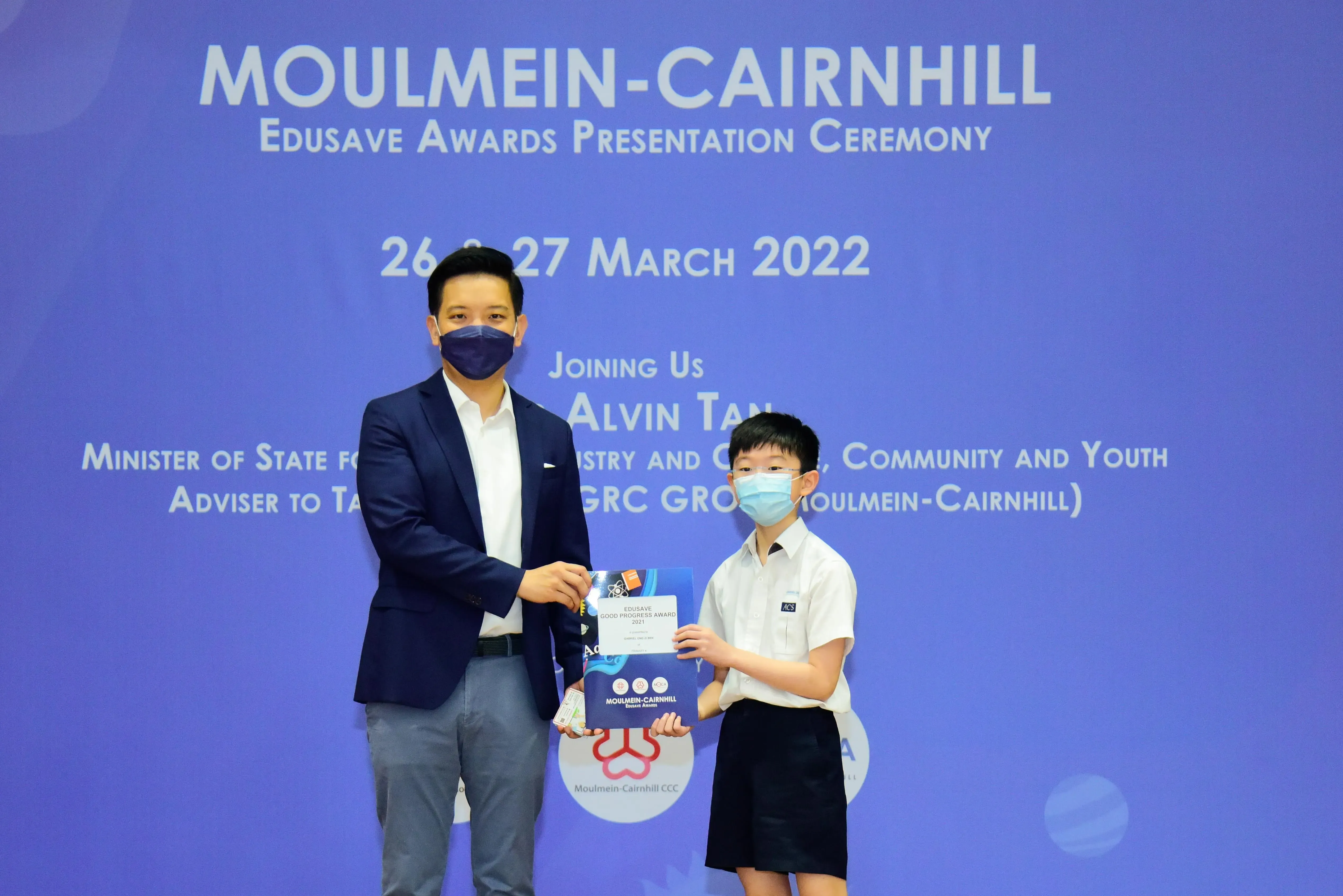 An adult in a suit and a young student in uniform, both masked, hold an award at the Moulmein-Cairnhill Edusave ceremony.