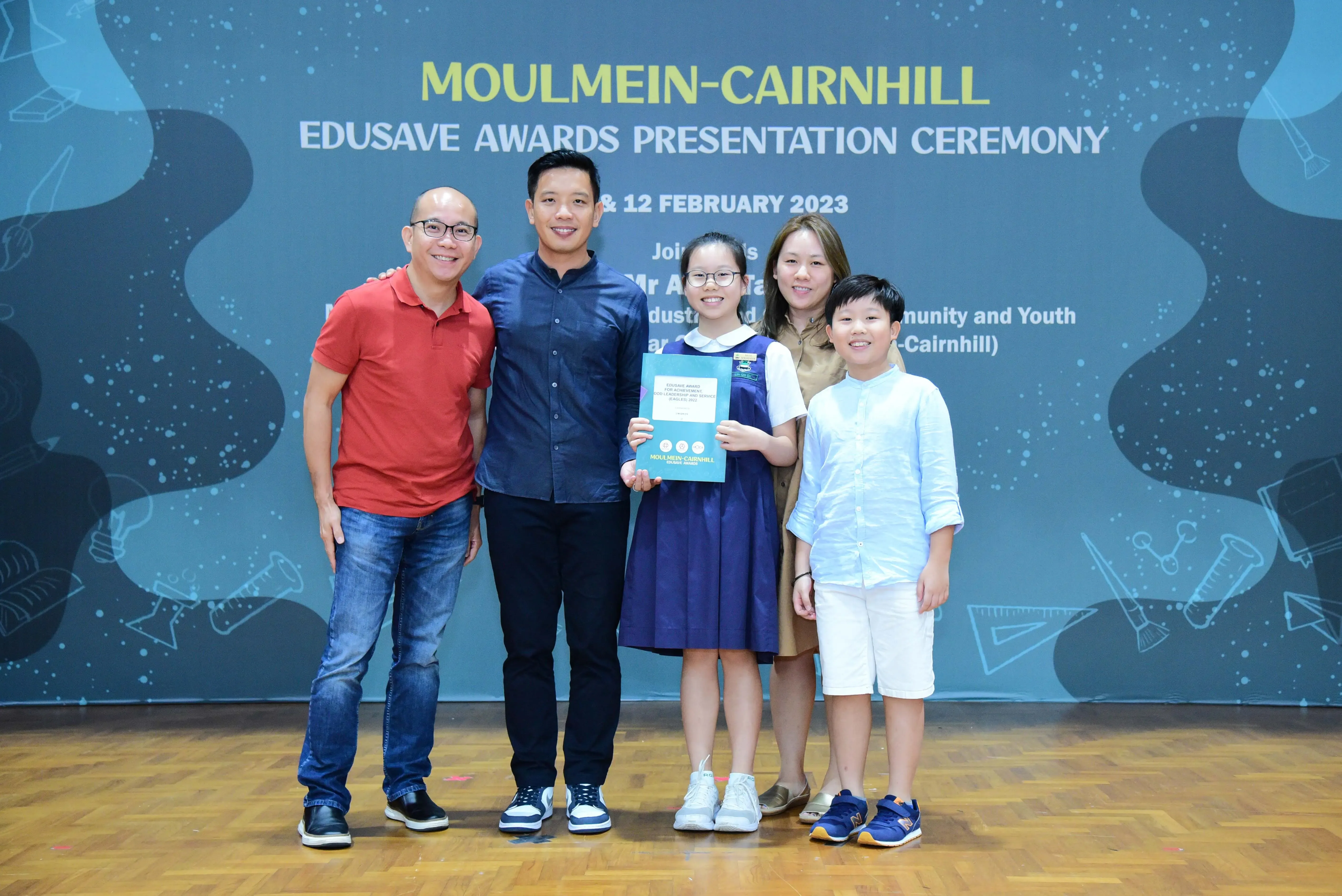 Five people pose on stage at the Moulmein-Cairnhill Edusave Awards Presentation Ceremony, with a girl holding a certificate.