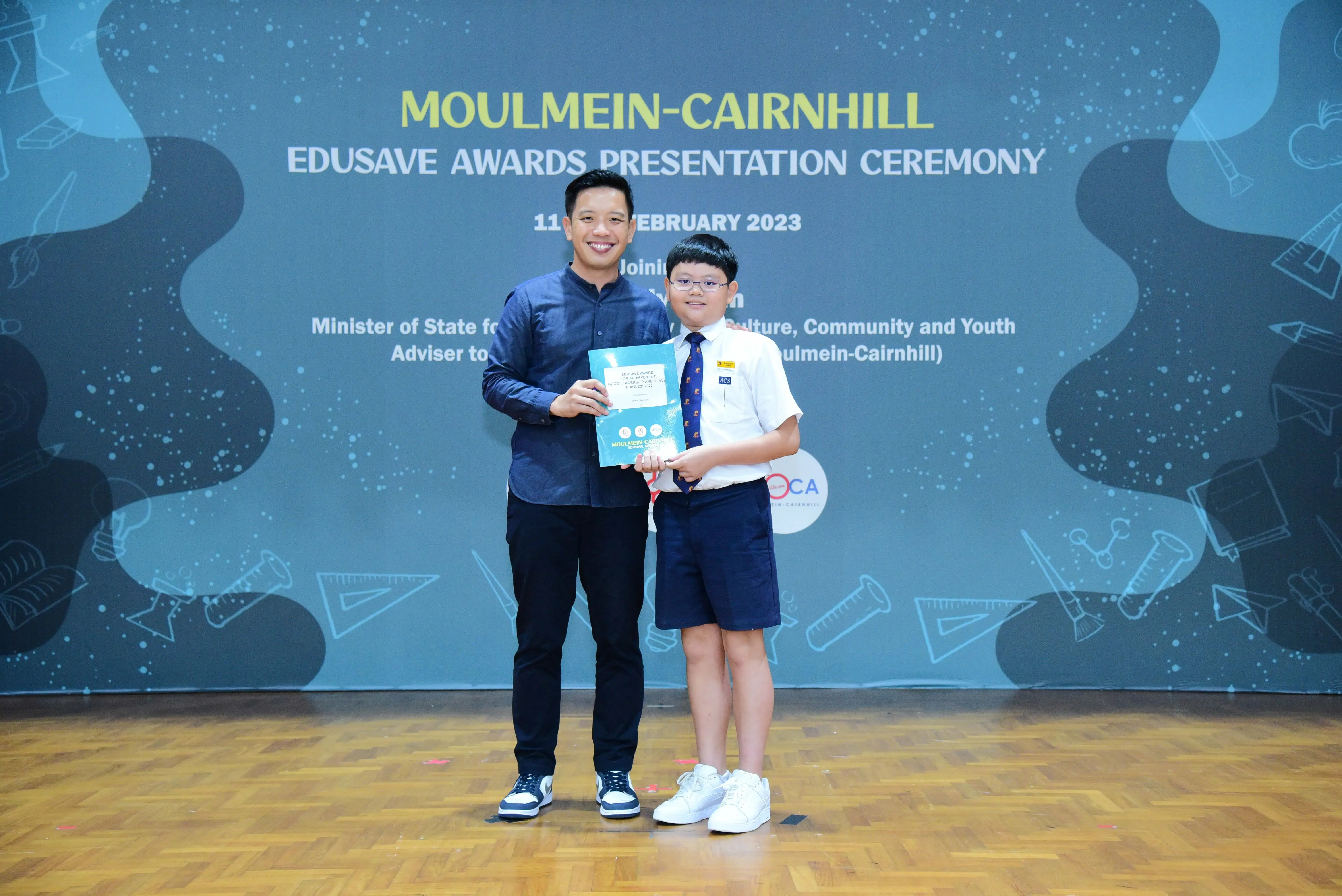 An adult man and a young boy in school uniform hold a certificate at the Moulmein-Cairnhill Edusave Awards Ceremony 2023.