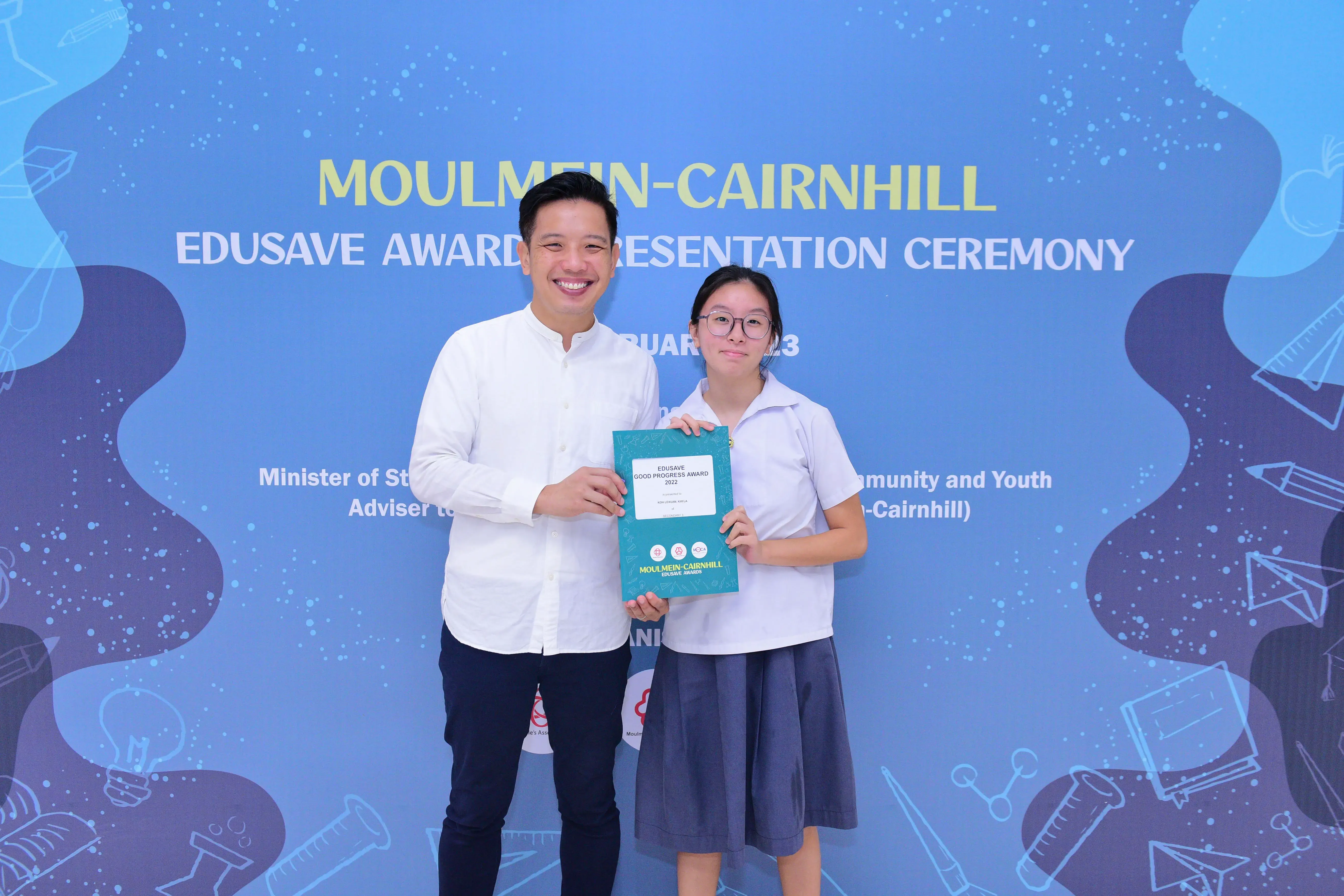 A man and a young girl hold a teal Edusave Good Progress Award certificate in front of a blue ceremony backdrop.