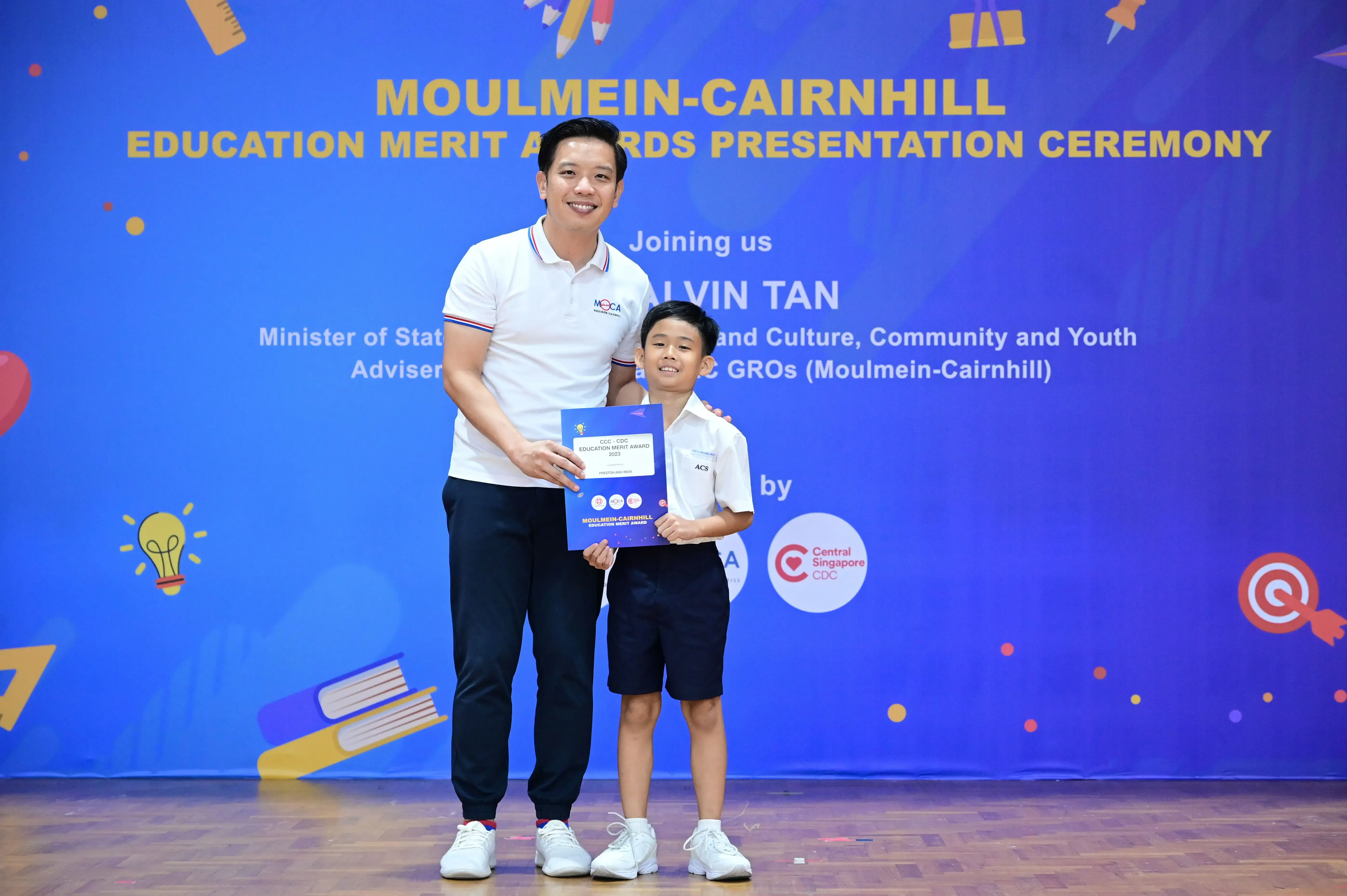 A man and a young boy holding a certificate at the Moulmein-Cairnhill Education Merit Awards ceremony.