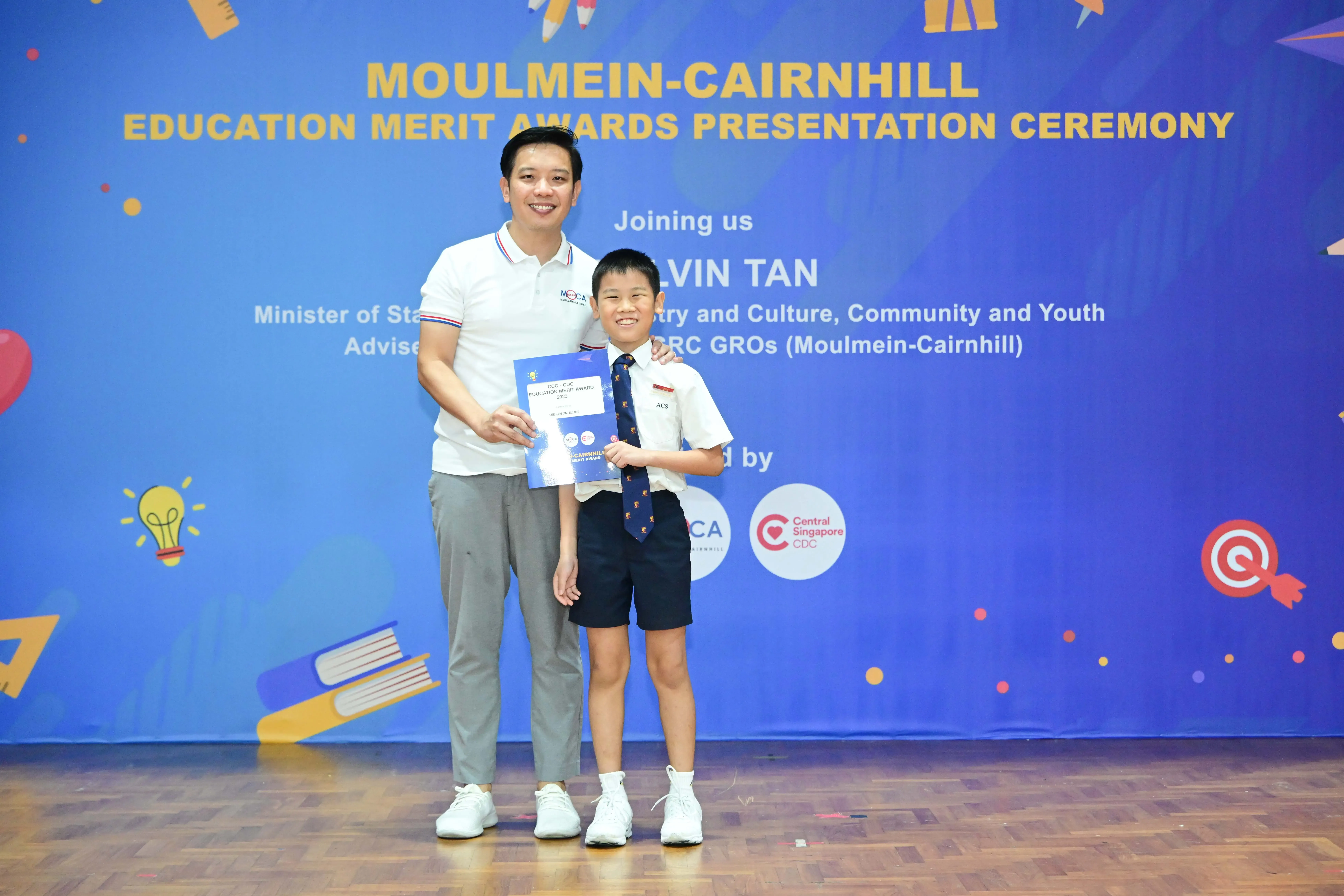 A man and a young boy in school uniform hold a certificate at the Moulmein-Cairnhill Education Merit Awards ceremony.