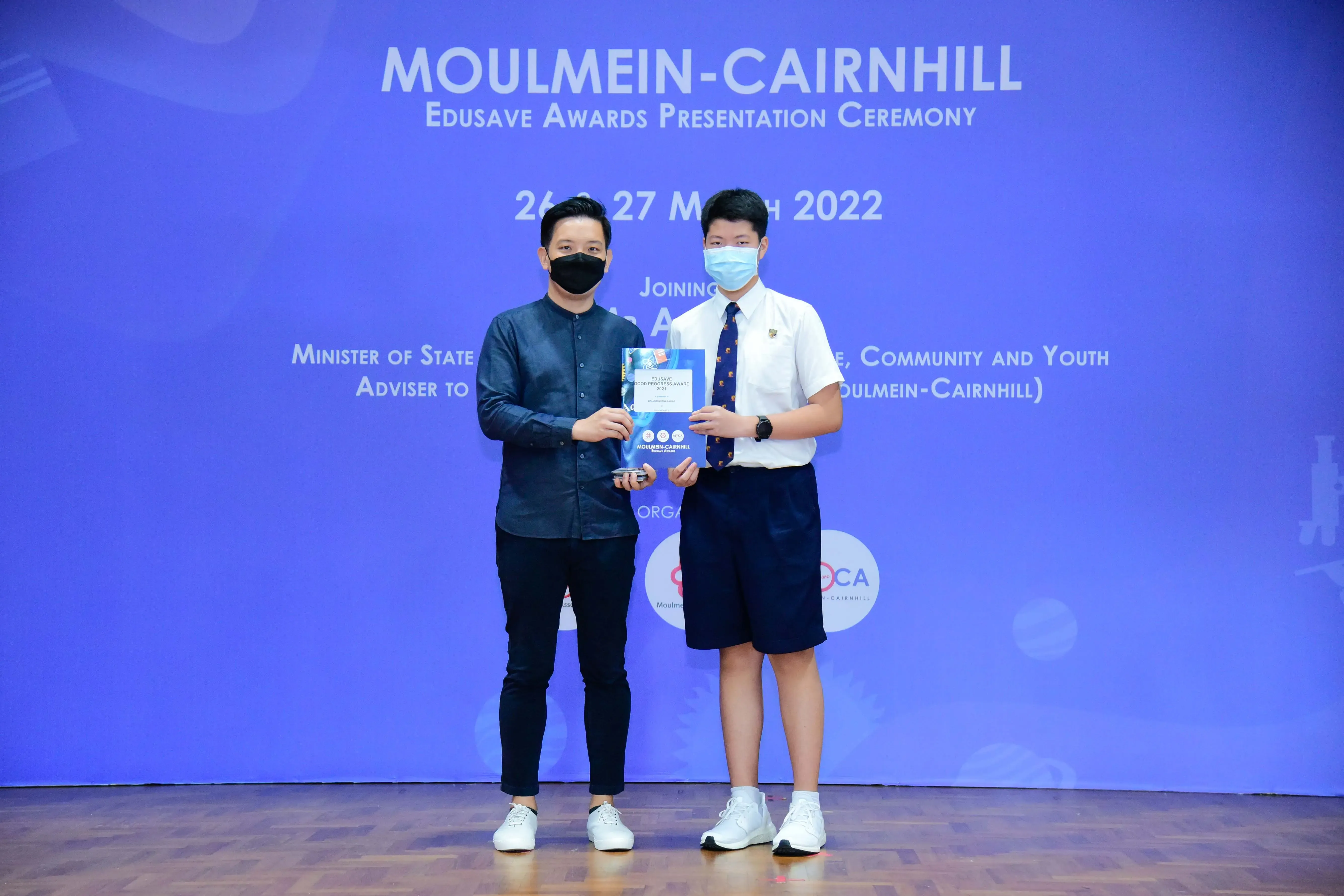 Two masked men hold an award at the Moulmein-Cairnhill Edusave Awards Presentation Ceremony, March 2022.