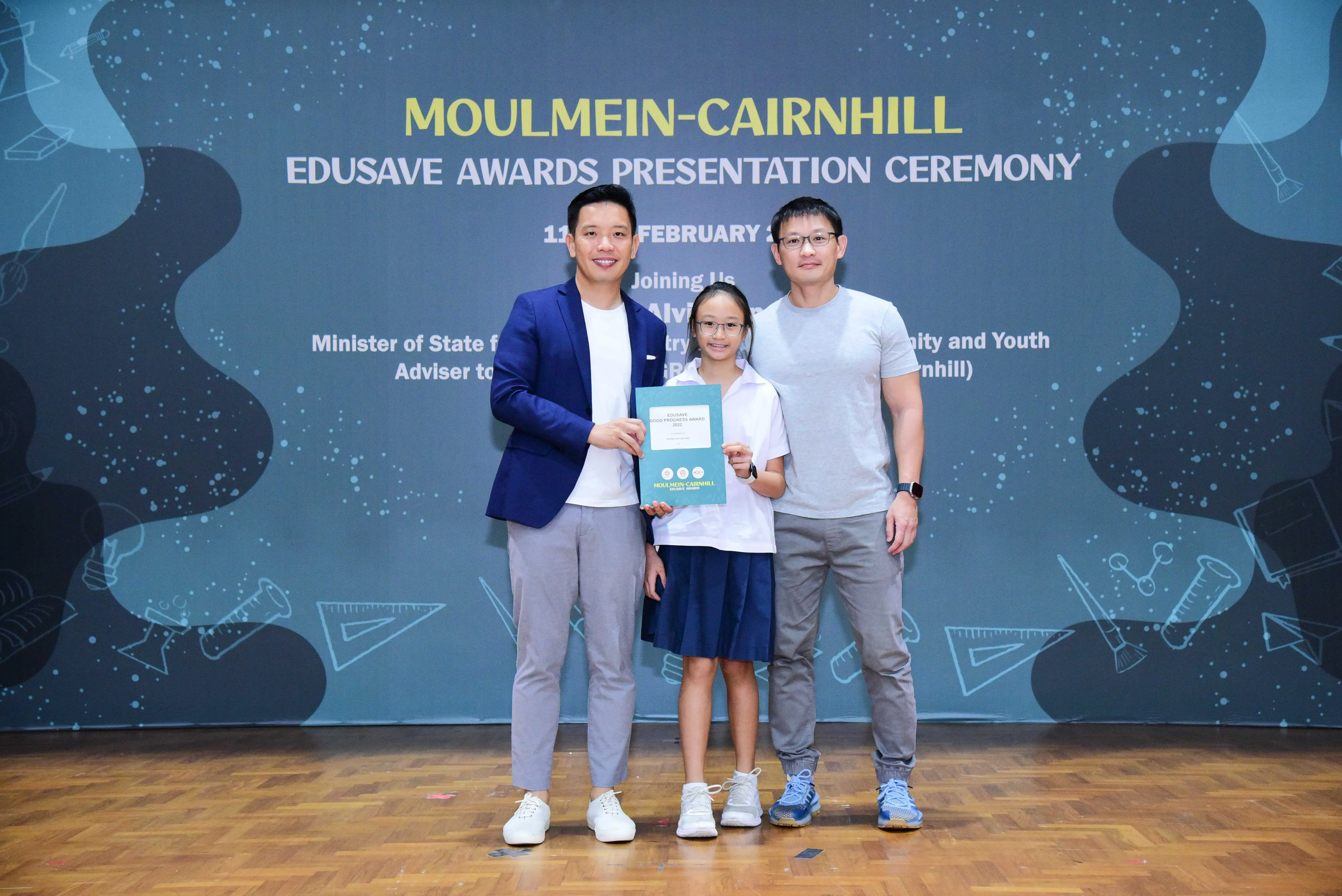 Two men and a young girl in school uniform hold an award at the Moulmein-Cairnhill Edusave Awards Presentation Ceremony.