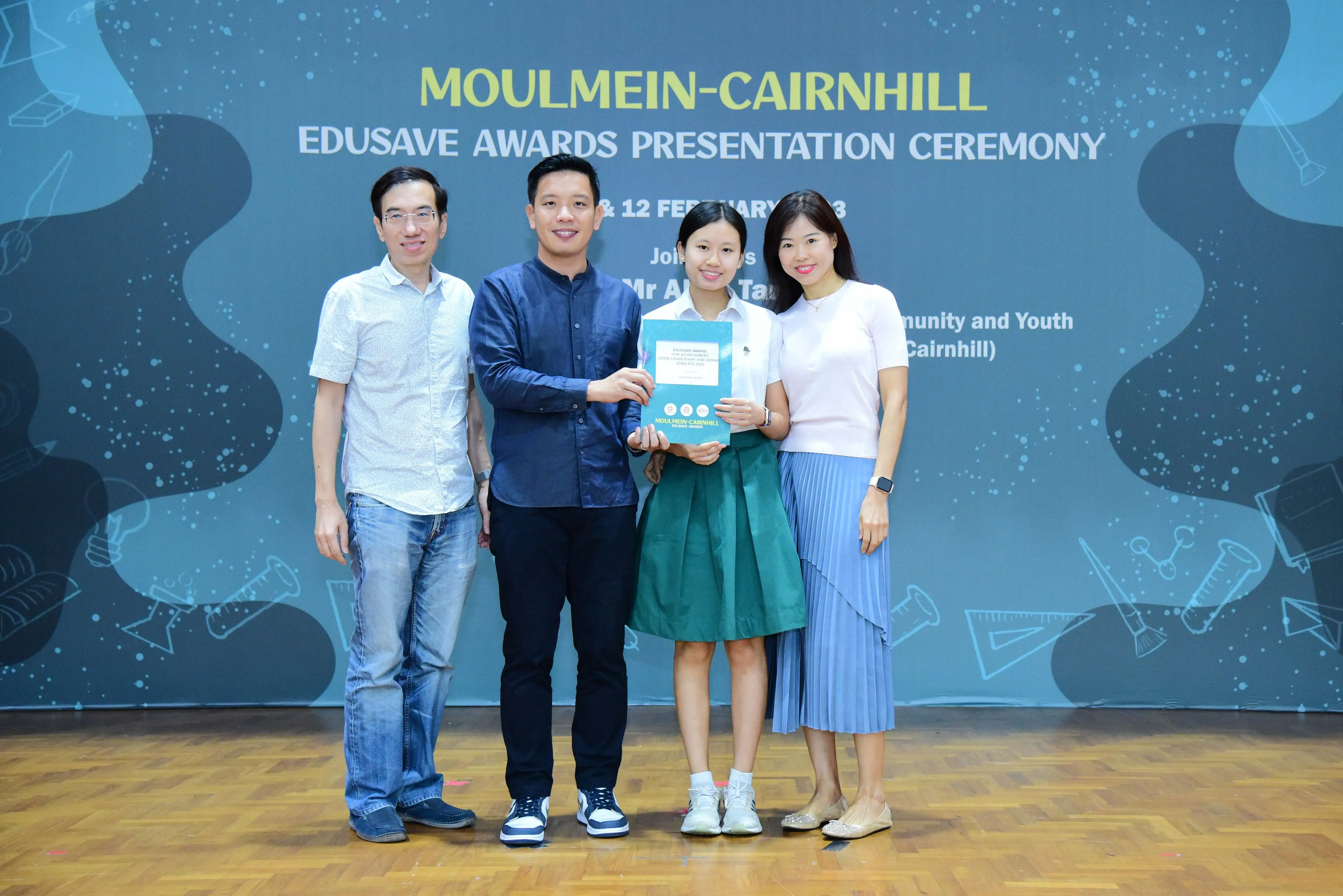 Four people pose at the Moulmein-Cairnhill Edusave Awards Presentation Ceremony, with a student holding an award booklet.