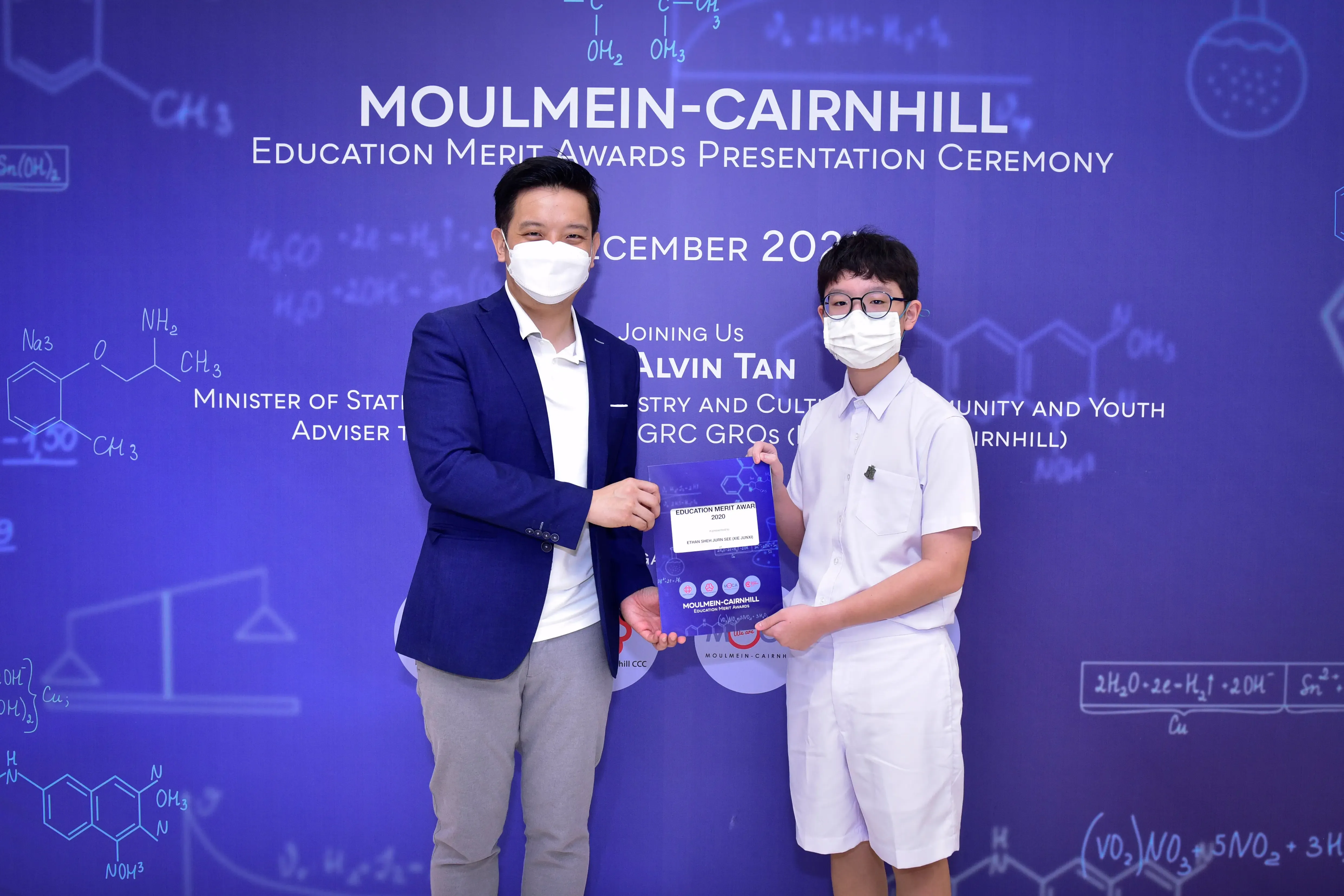Two masked individuals hold an award certificate at the Moulmein-Cairnhill Education Merit Awards Presentation Ceremony.