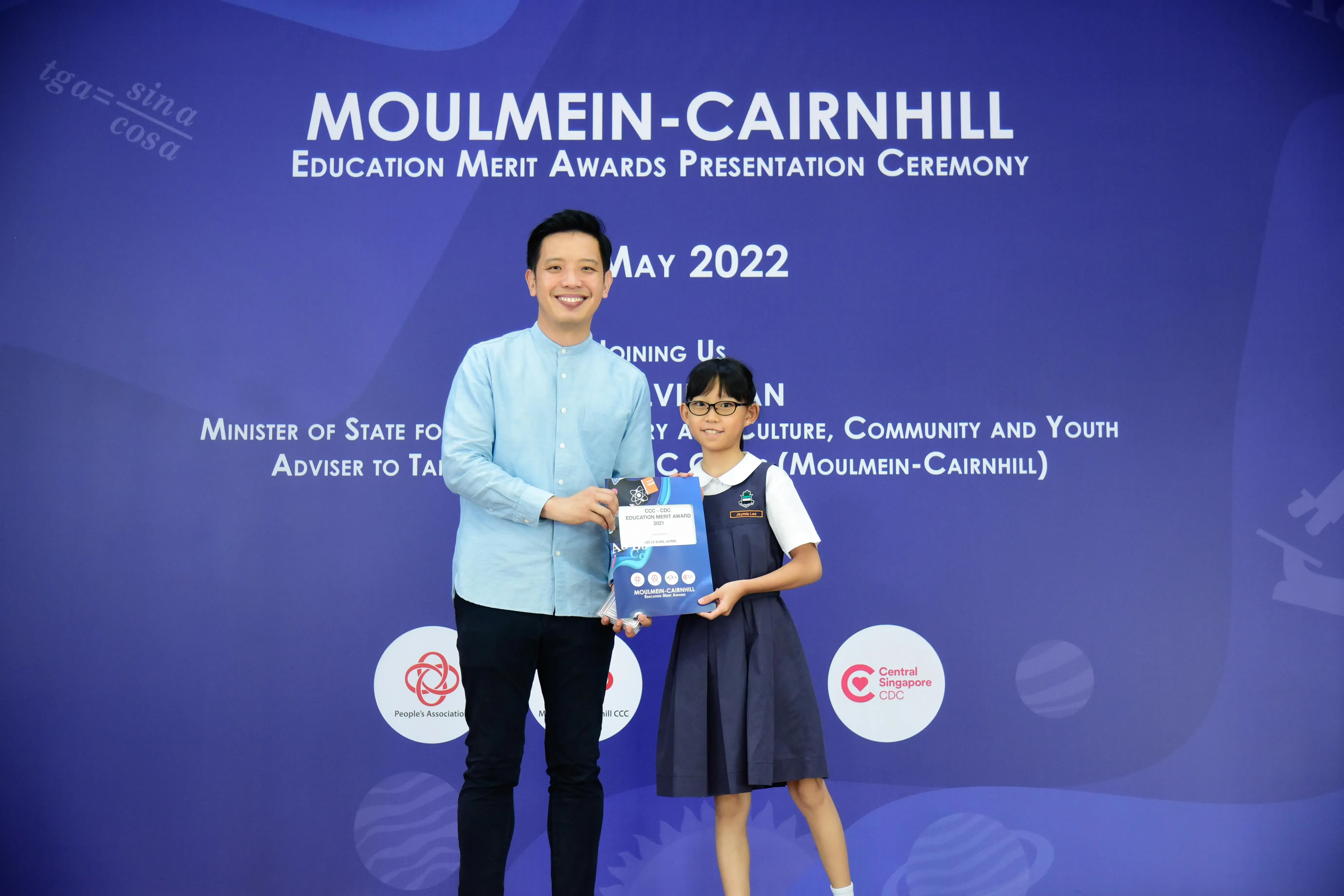 A man and a young girl in school uniform hold an award together at the Moulmein-Cairnhill Education Merit Awards, May 2022.